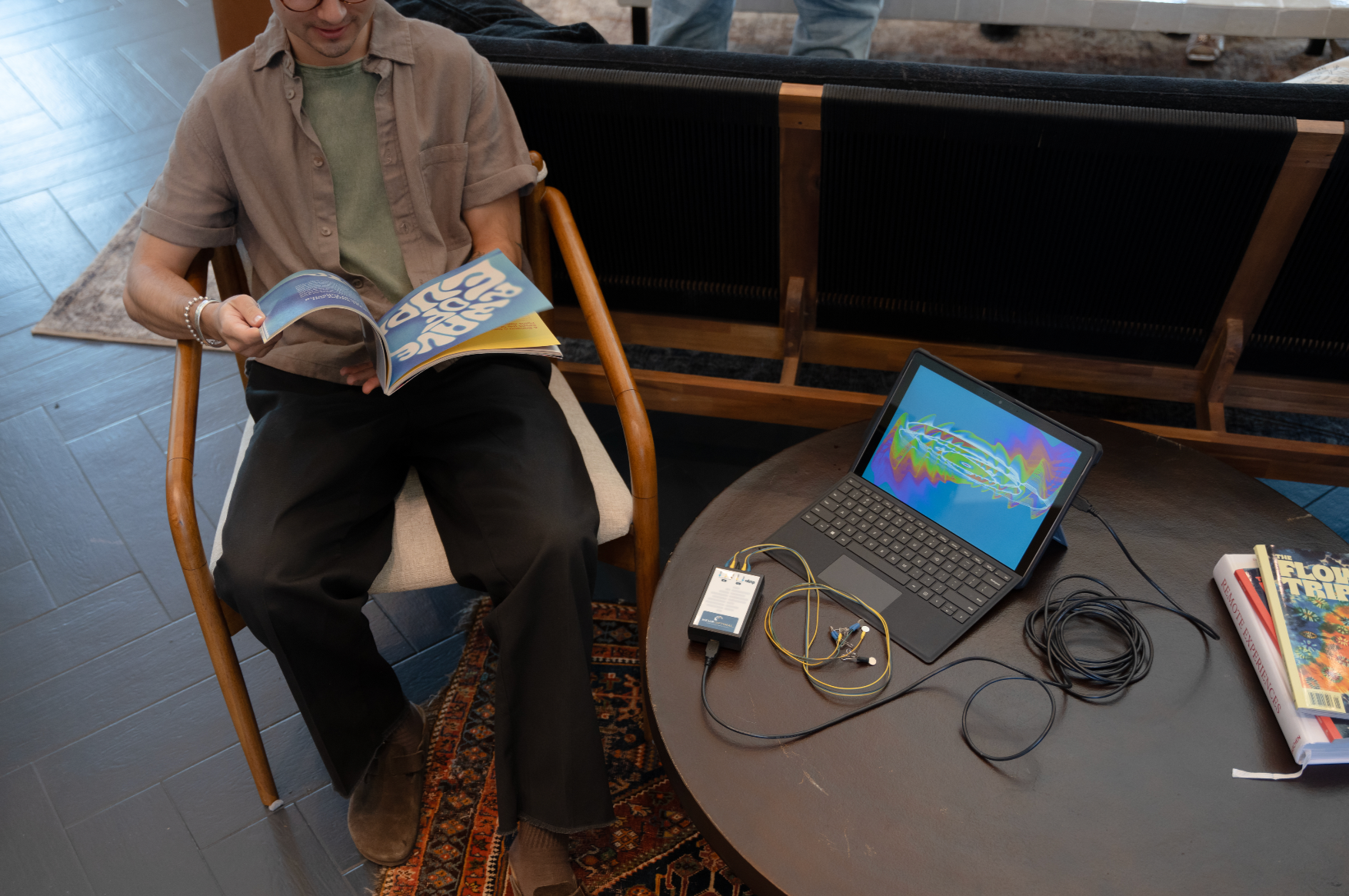 Person sitting in a wooden chair reading a magazine next to a table with a laptop, connected electronic device, and books.