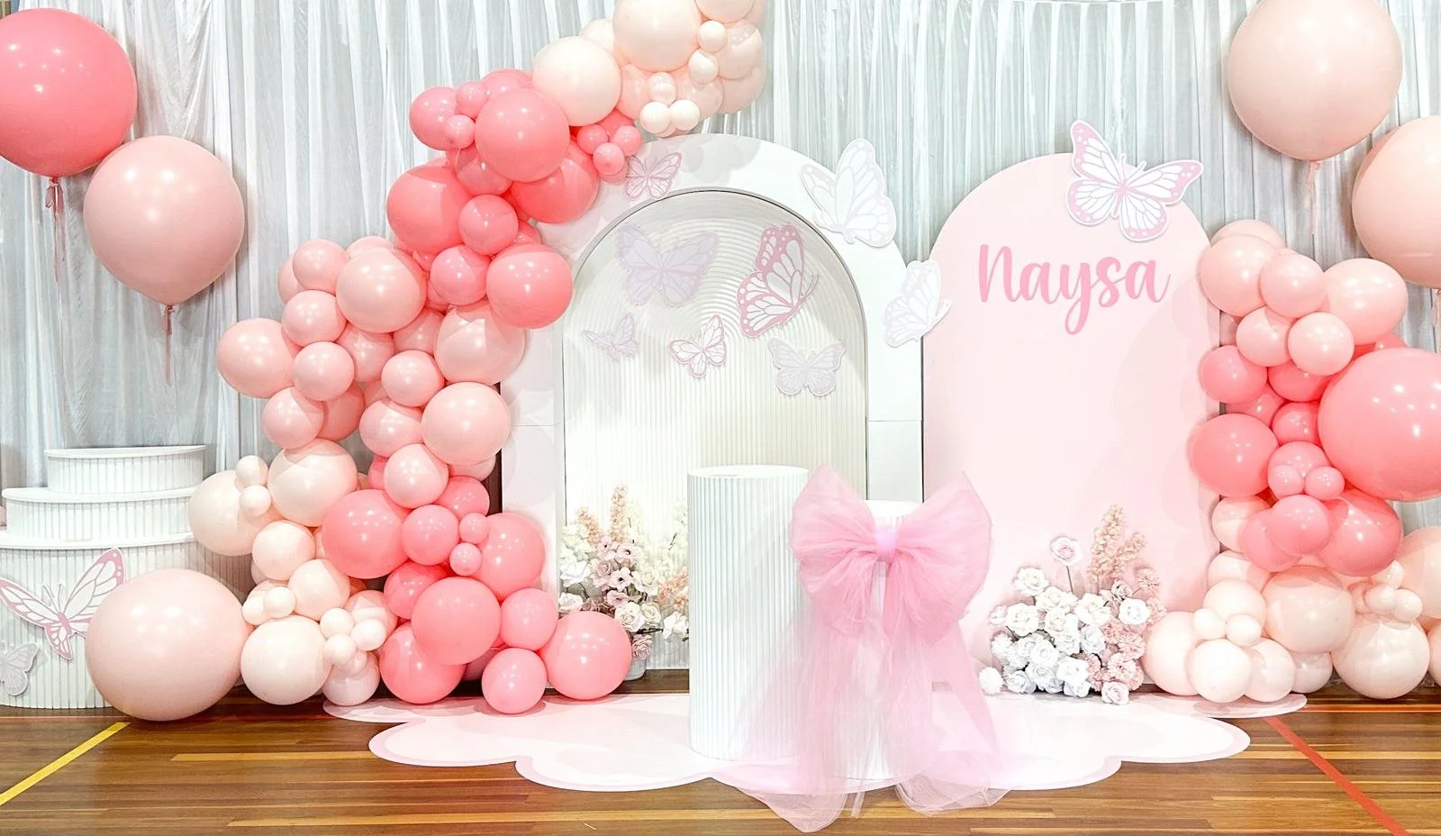 Pink and white balloon decorations with flowers, butterflies, and a pink sign that says "Naysa" for a celebration backdrop.
