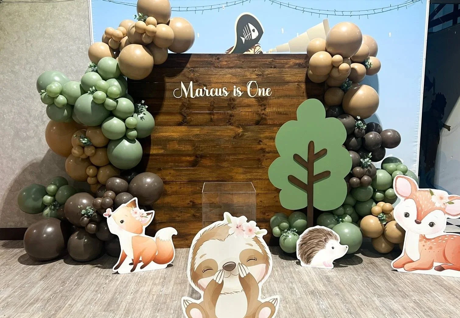 Decorative backdrop with balloons and cutouts for a child's first birthday party, featuring a wooden sign that reads "Marcus is One," cartoon animals, and a tree.