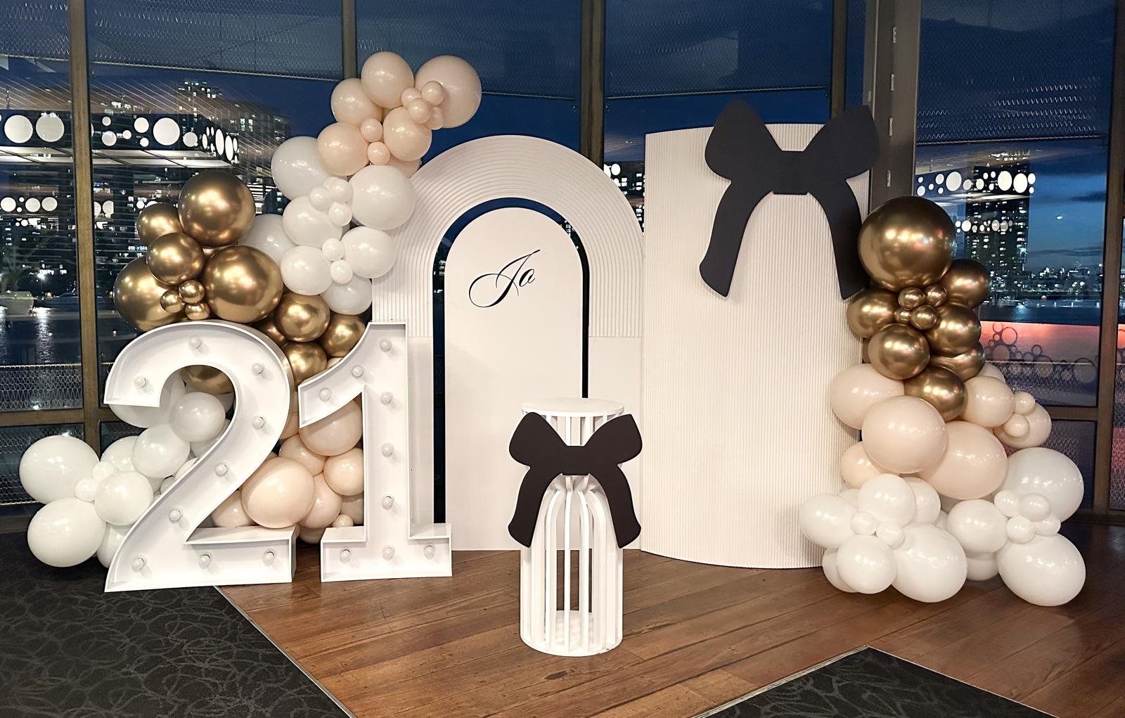 Decorative celebration setup with white and gold balloons, large marquee numbers '21', black bow decorations, and a white backdrop with the word 'Jo' written on it, titled for a 21st birthday.