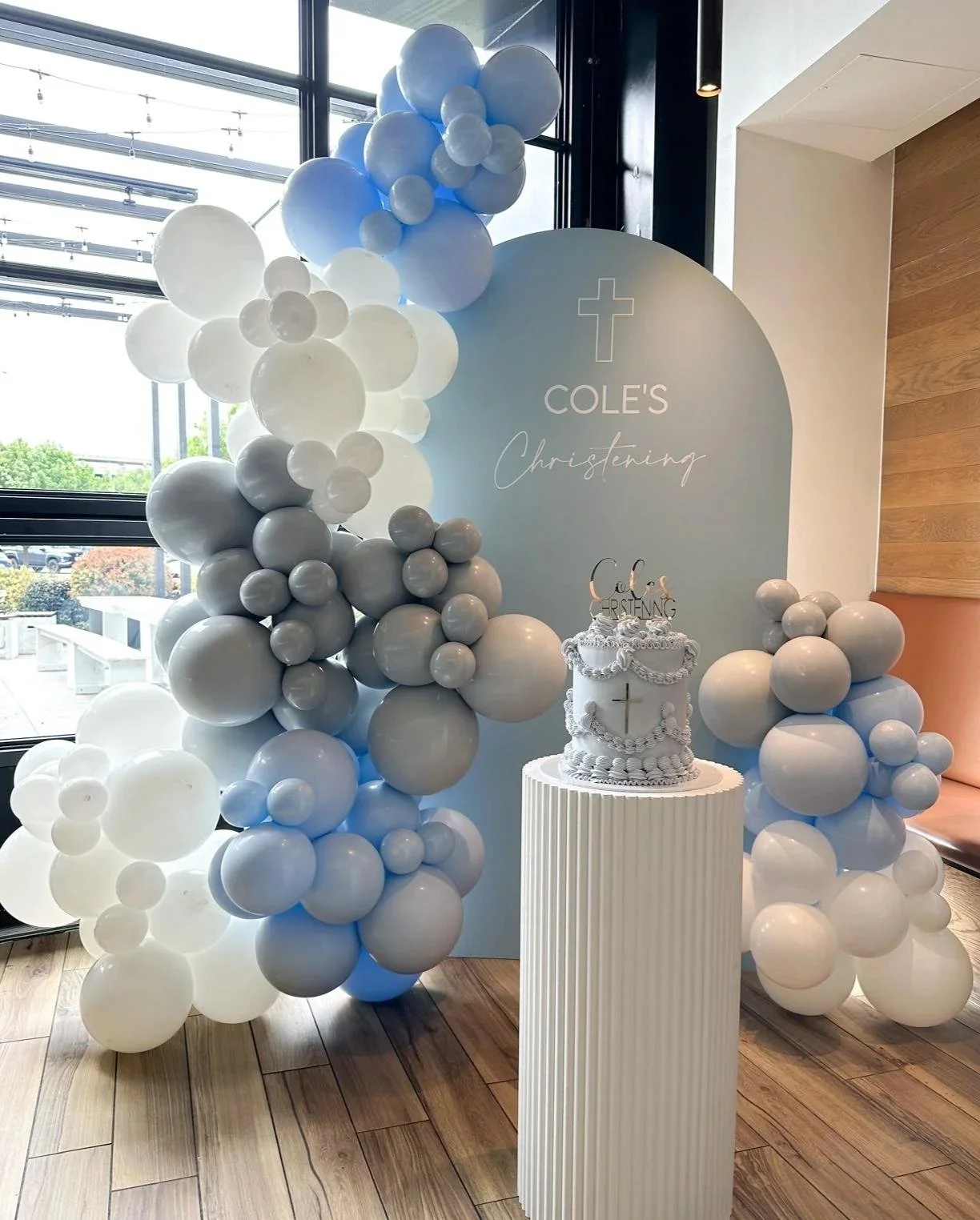 A baptism or christening decoration setup featuring a large circular backdrop with a cross and the words 'Cole's Christening,' surrounded by balloon arrangements in shades of white, gray, and light blue. There is a decorated cake on a white pedestal table in front of the backdrop.