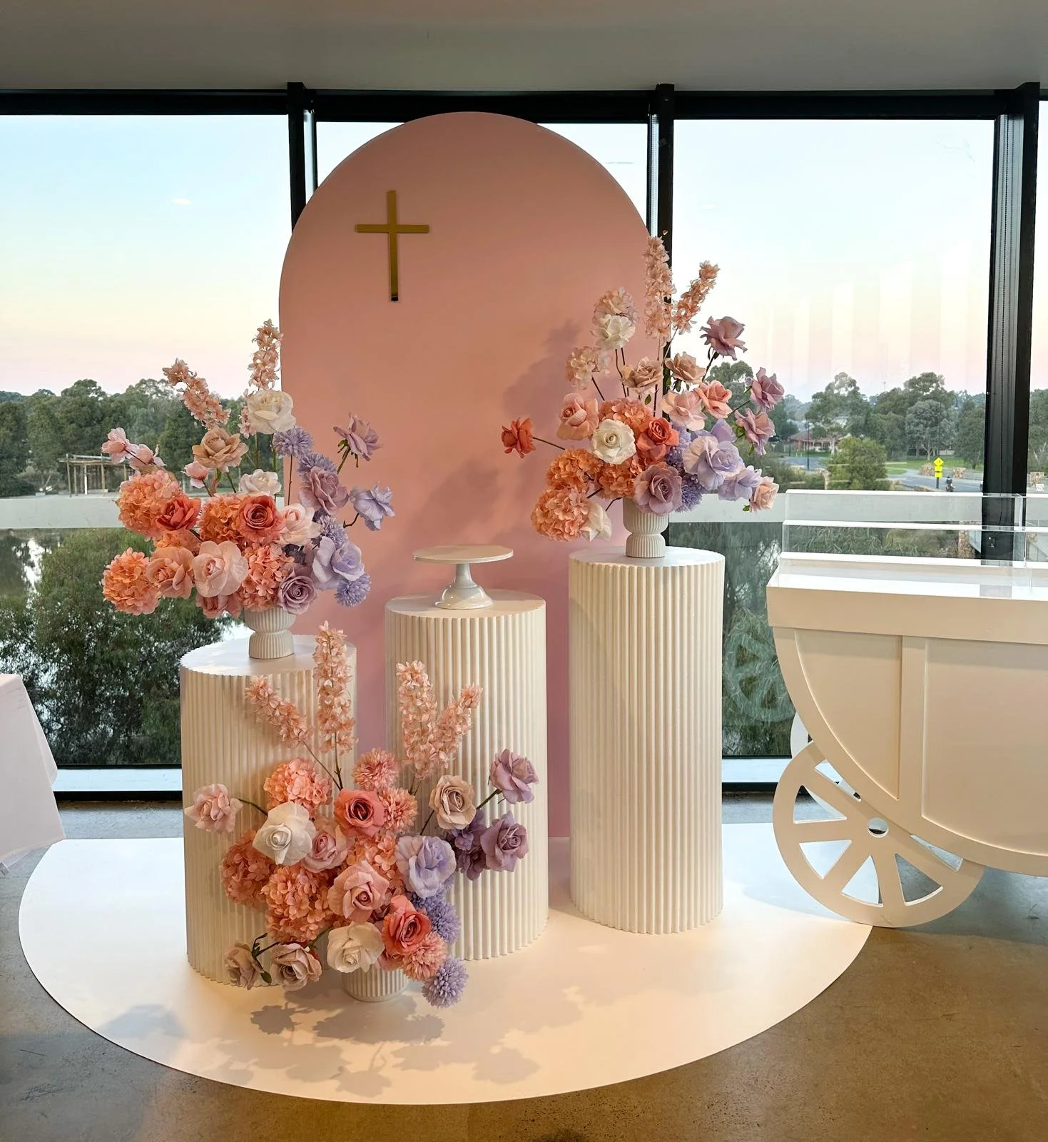Decorative display with pink and purple flowers, white pedestals, a pink backdrop with a gold cross, and a white cart, set in front of large windows showing a tree-lined landscape.