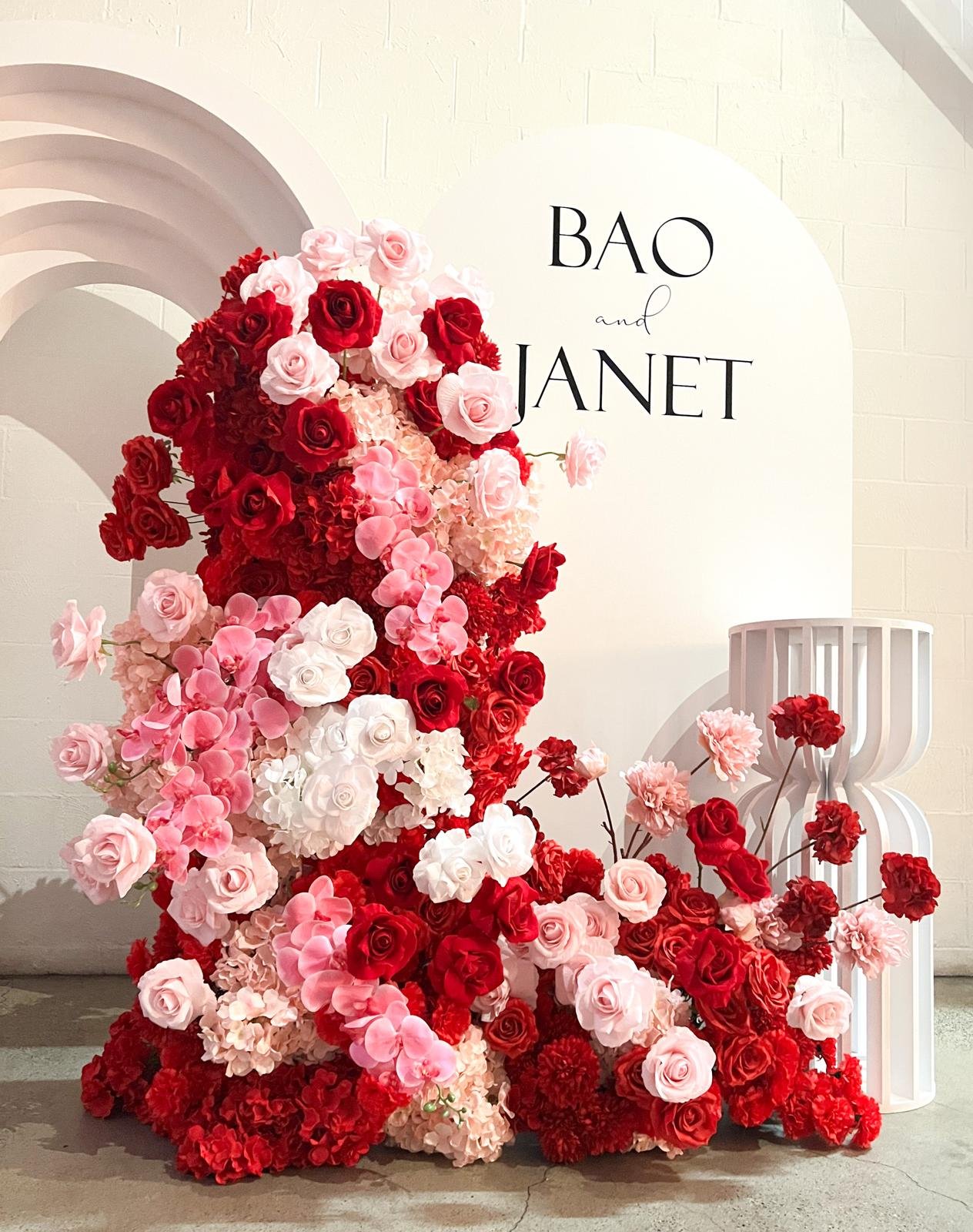A floral arrangement with red, pink, and white roses, hydrangeas, and carnations next to a white decorative sculpture with vertical slats, placed in front of a white wall with the names 'BAO and JANET' on a round white backdrop.