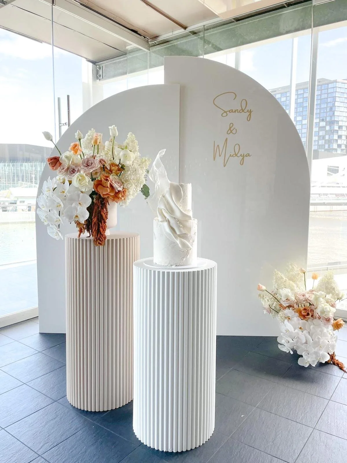 A white event display with a floral arrangement, a white sculptural cake, and a backdrop with gold cursive writing that reads "Sandy & Midya". The setup is indoors near large windows with a cityscape view.