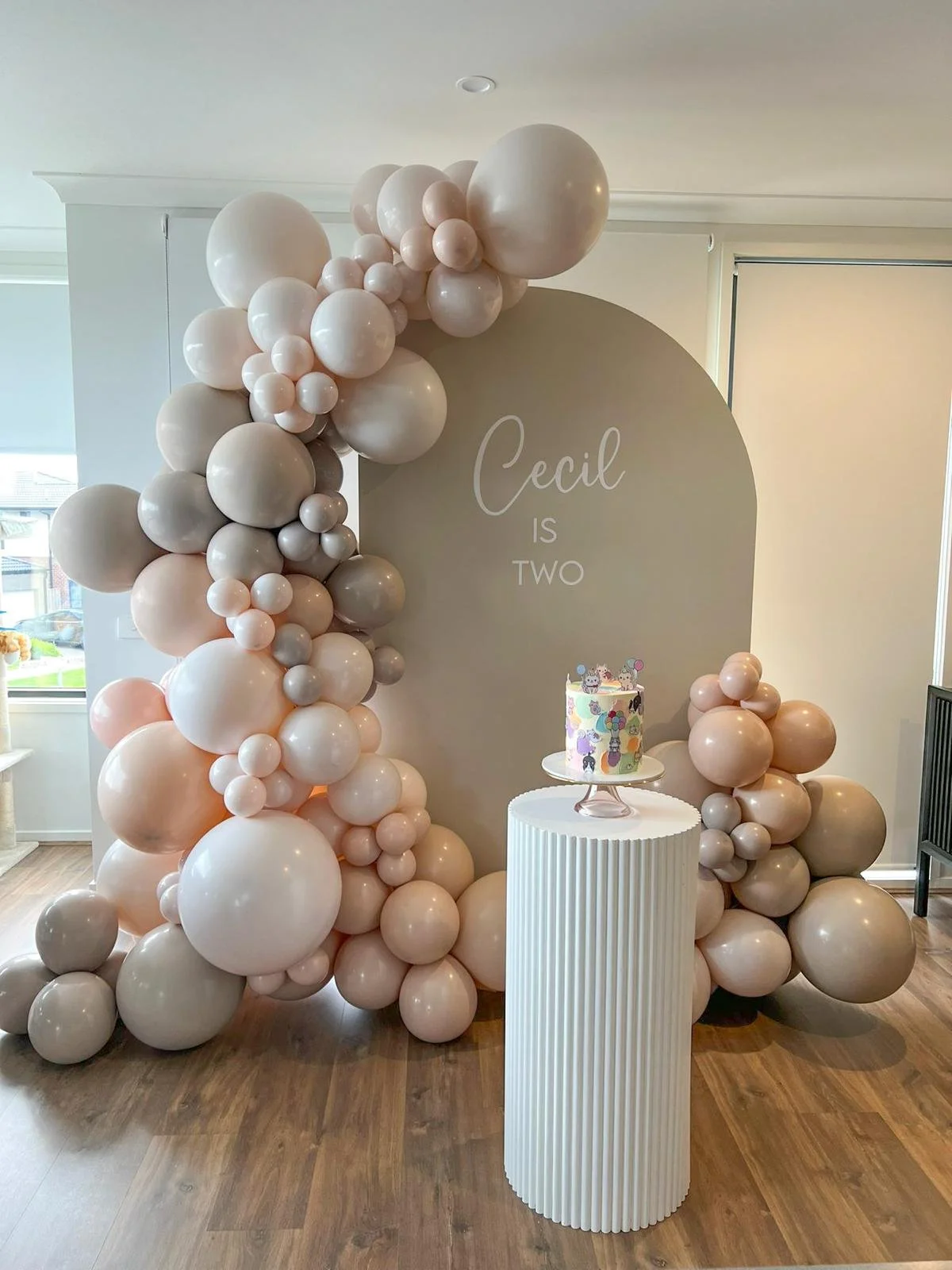 Decorative birthday setup with an arch of pink, white, and beige balloons, a cake on a white-tubed pedestal, a background sign reading 'Cecil is Two,' and a colorful cartoon-themed cake with animals.