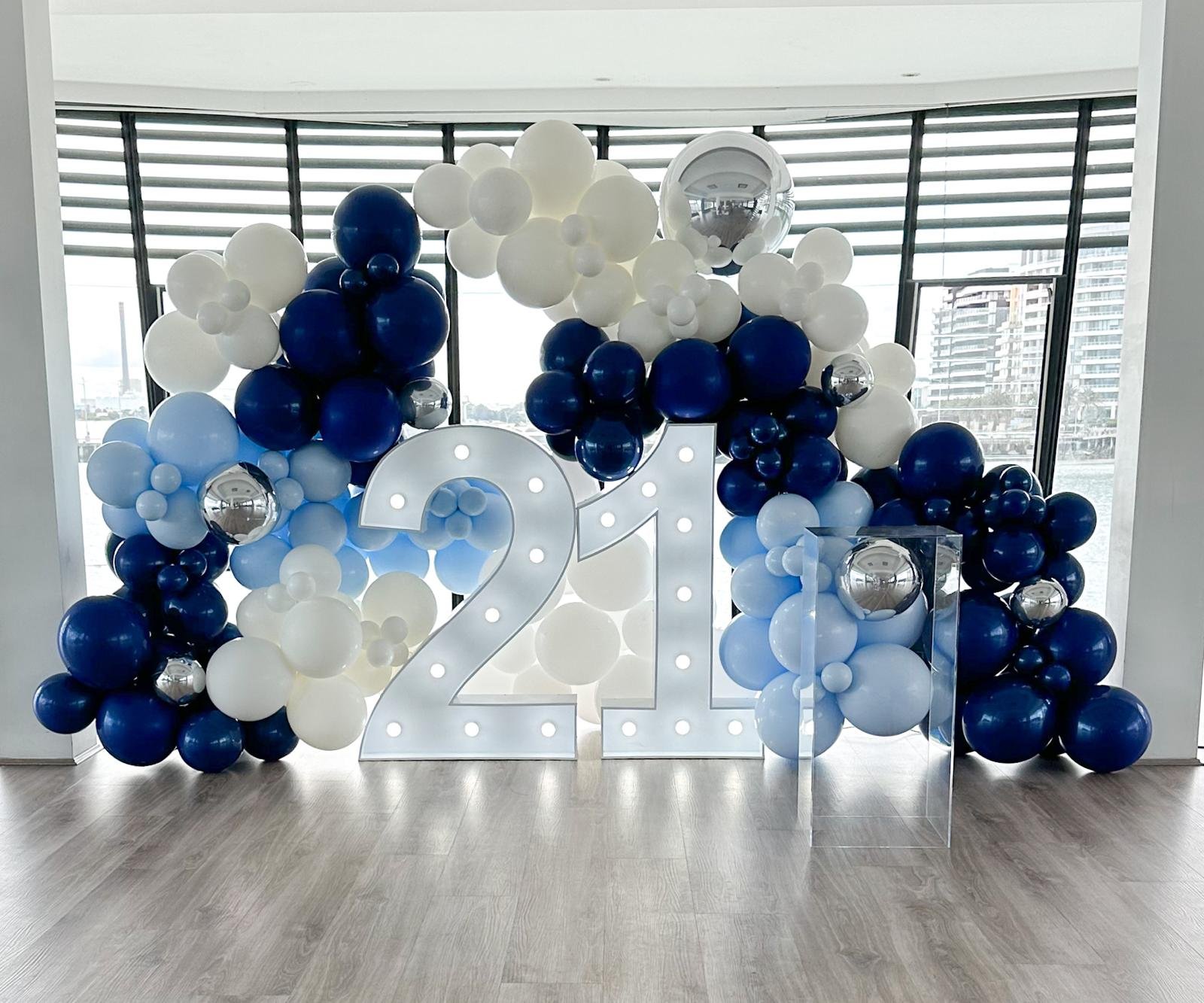 Balloon backdrop with white, light blue, dark blue, and silver balloons, large illuminated number 21, and a clear acrylic stand in a modern room with windows showing city buildings.