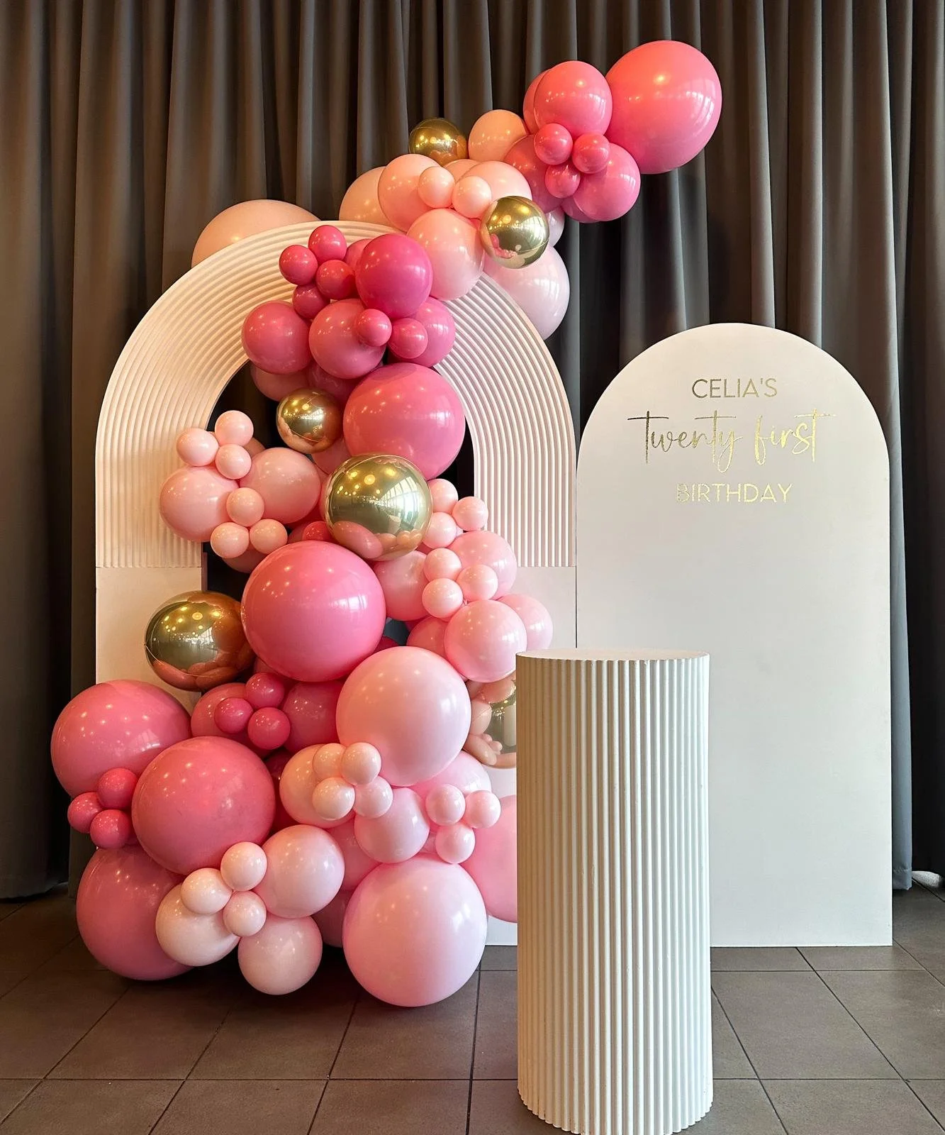 A birthday decoration backdrop featuring a large pink balloon arrangement with gold and lighter pink balloons, positioned next to a white arched sign and a ripple plint