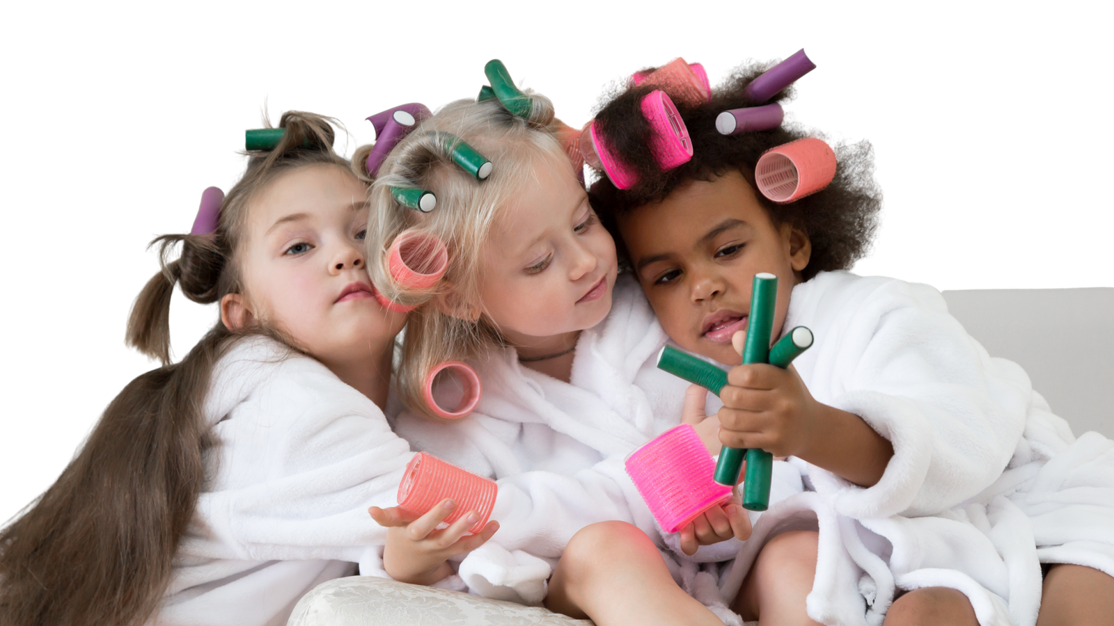Three young girls in plush terrycloth robes and hair rollers in their hair, sitting closely together, engaging with hair curlers in a playful manner.