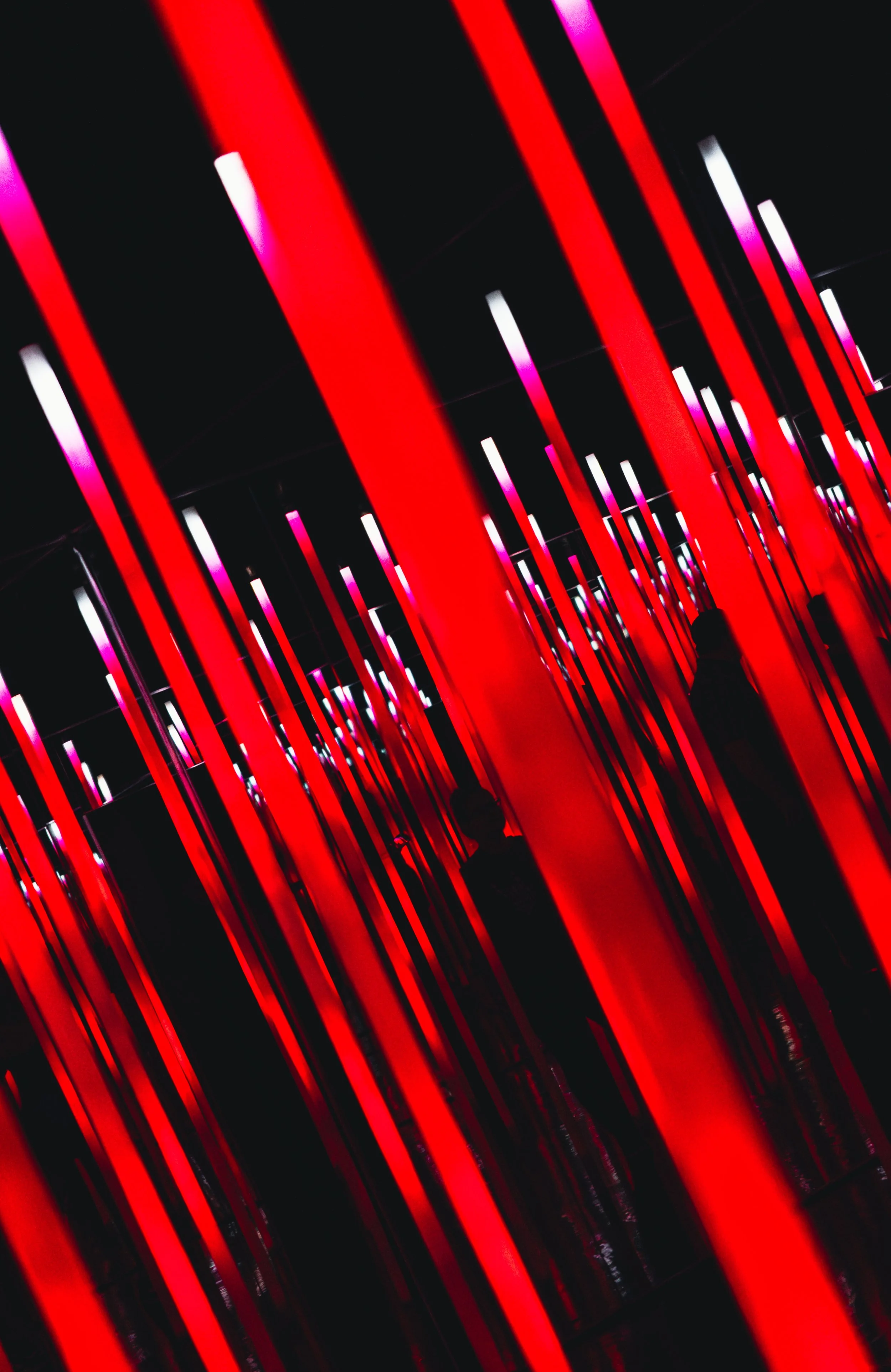 An immersive art installation featuring vertical, illuminated red and pink light bars creating a corridor effect, with silhouettes of people walking through.