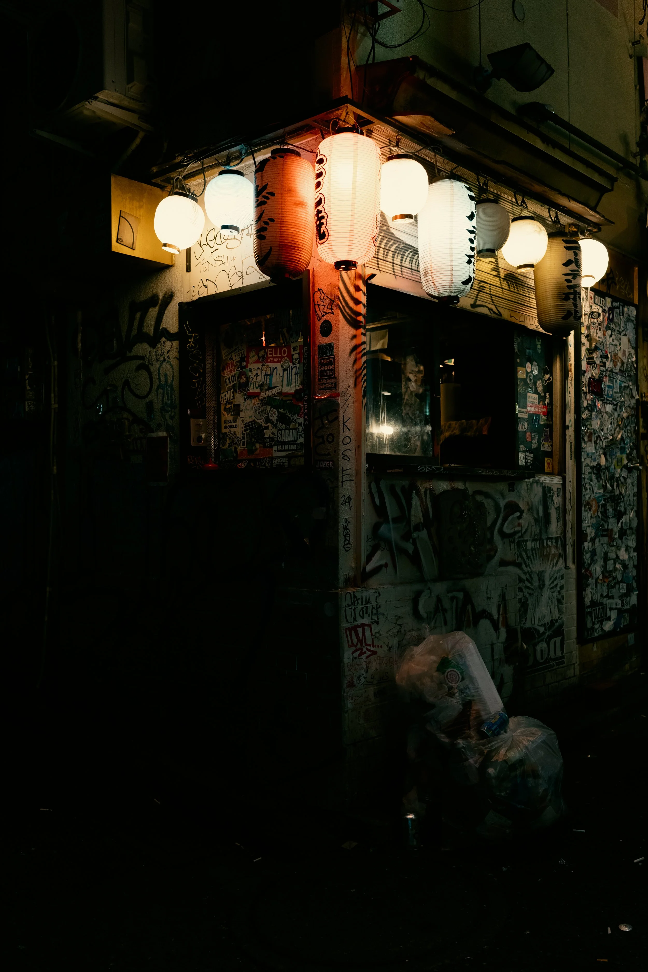 Nighttime street scene featuring a small street stall or booth illuminated by hanging Asian paper lanterns, with graffiti-covered walls and graffiti-covered shutter panels, and garbage bags on the sidewalk.