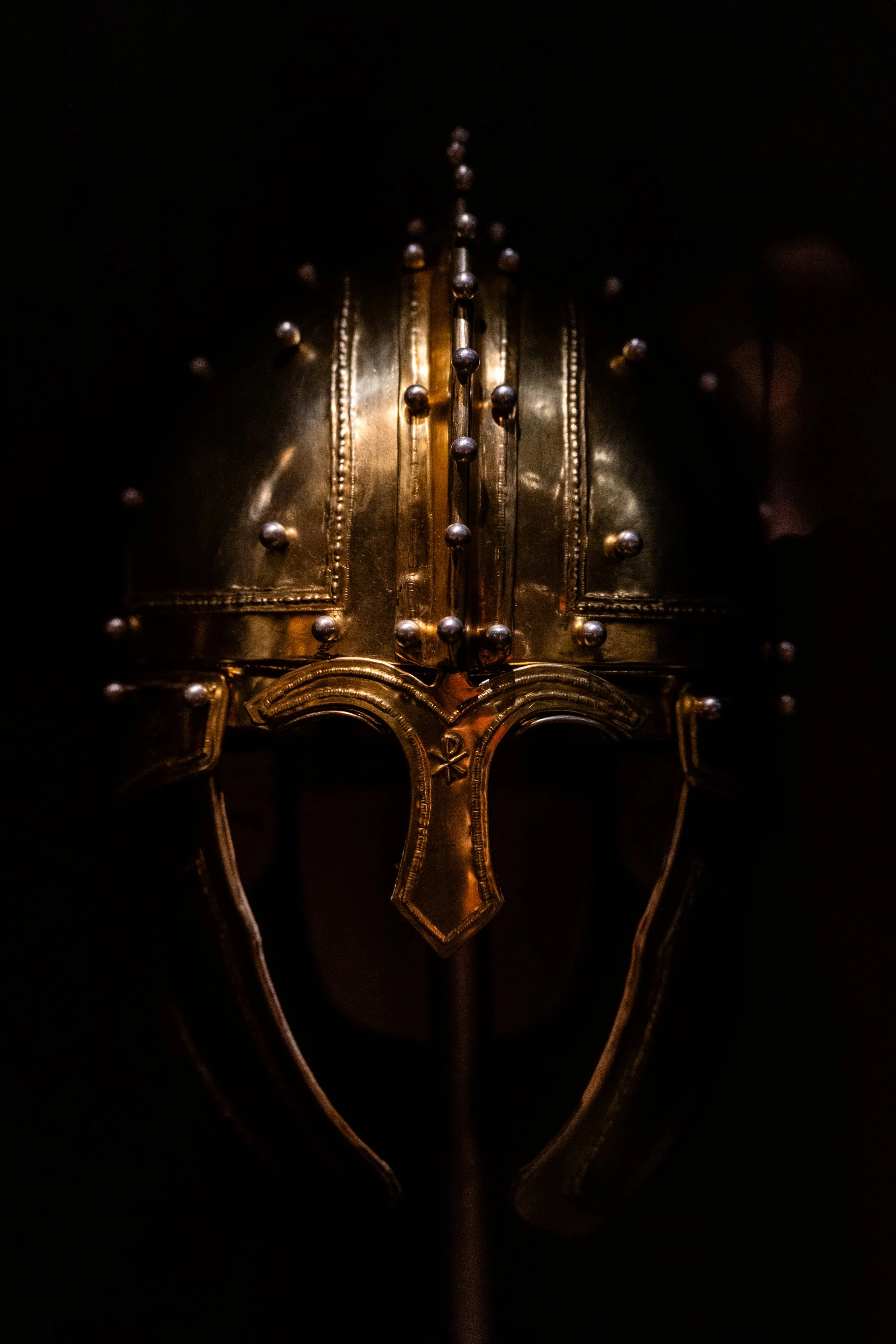A close-up of an ancient, ornate metallic helmet with decorative studs, displayed against a dark background.