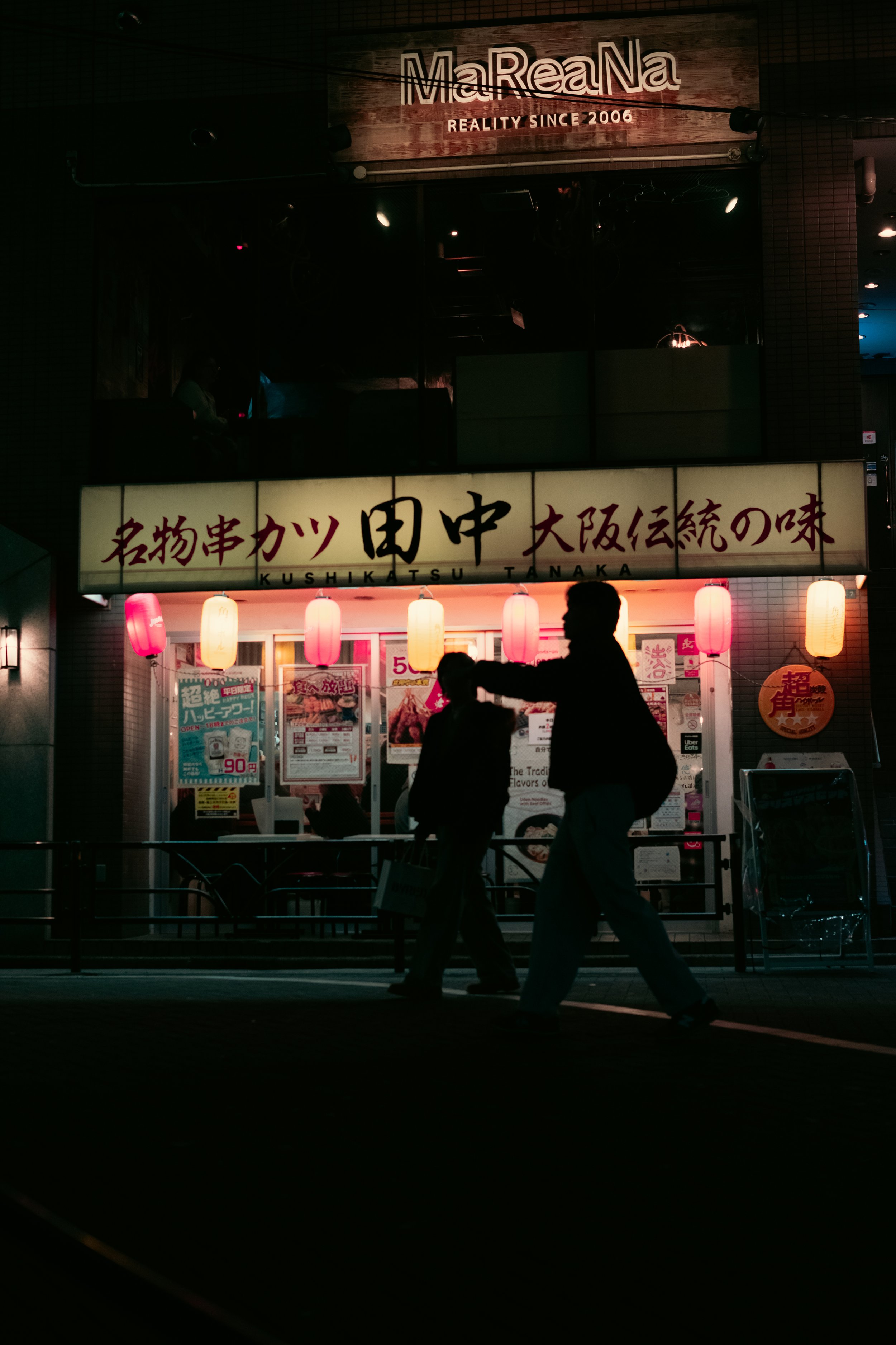 Nighttime Japanese street scene with two silhouetted people walking past a restaurant with illuminated lanterns and Japanese signage.