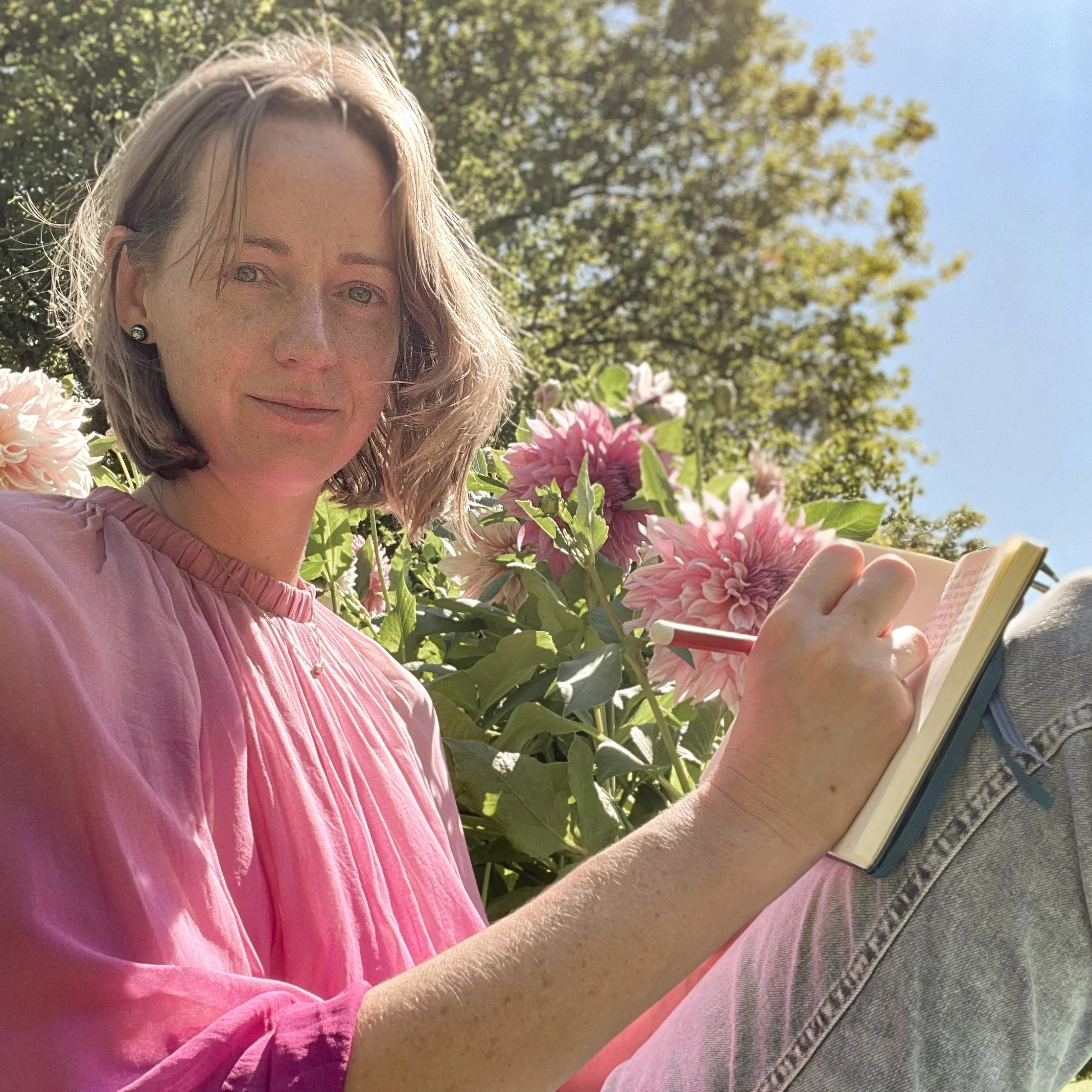 A woman with shoulder-length blonde hair wearing a pink shirt, holding a smartphone, standing outdoors among pink flowers and green trees under a clear blue sky.