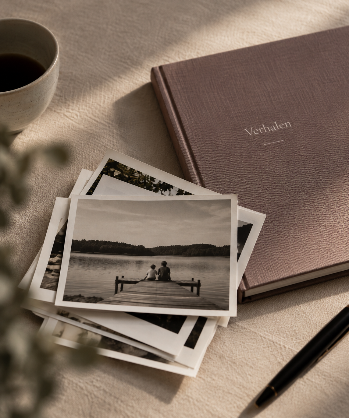 A table with a cup of coffee, a closed photo album labeled 'Verhalen', four black-and-white photographs of a lakeside scene with two children sitting by the water, and a black pen, with sunlight creating a warm, cozy atmosphere.