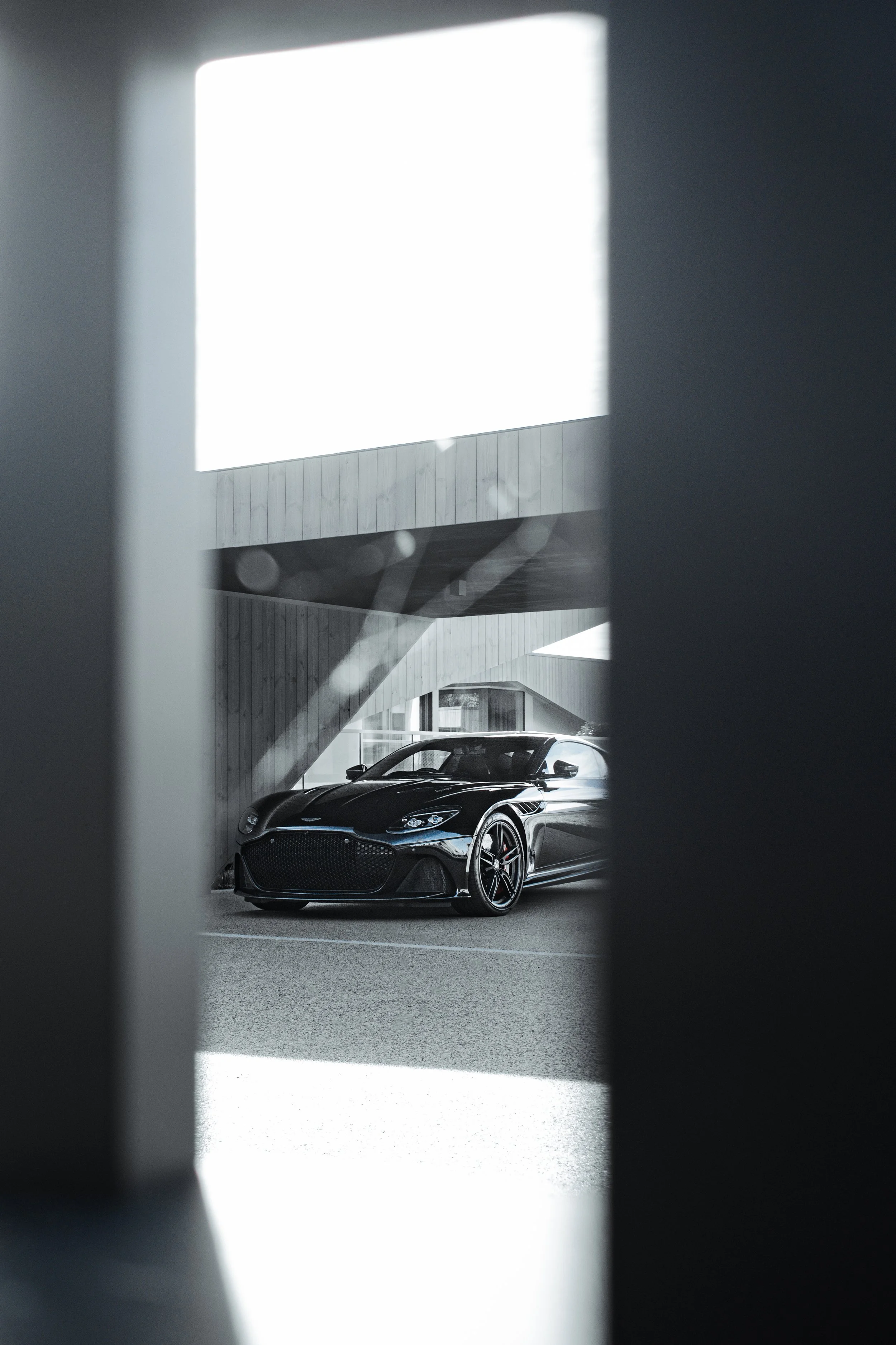 A black luxury sports car parked outdoors, viewed through a narrow gap between two vertical structures.