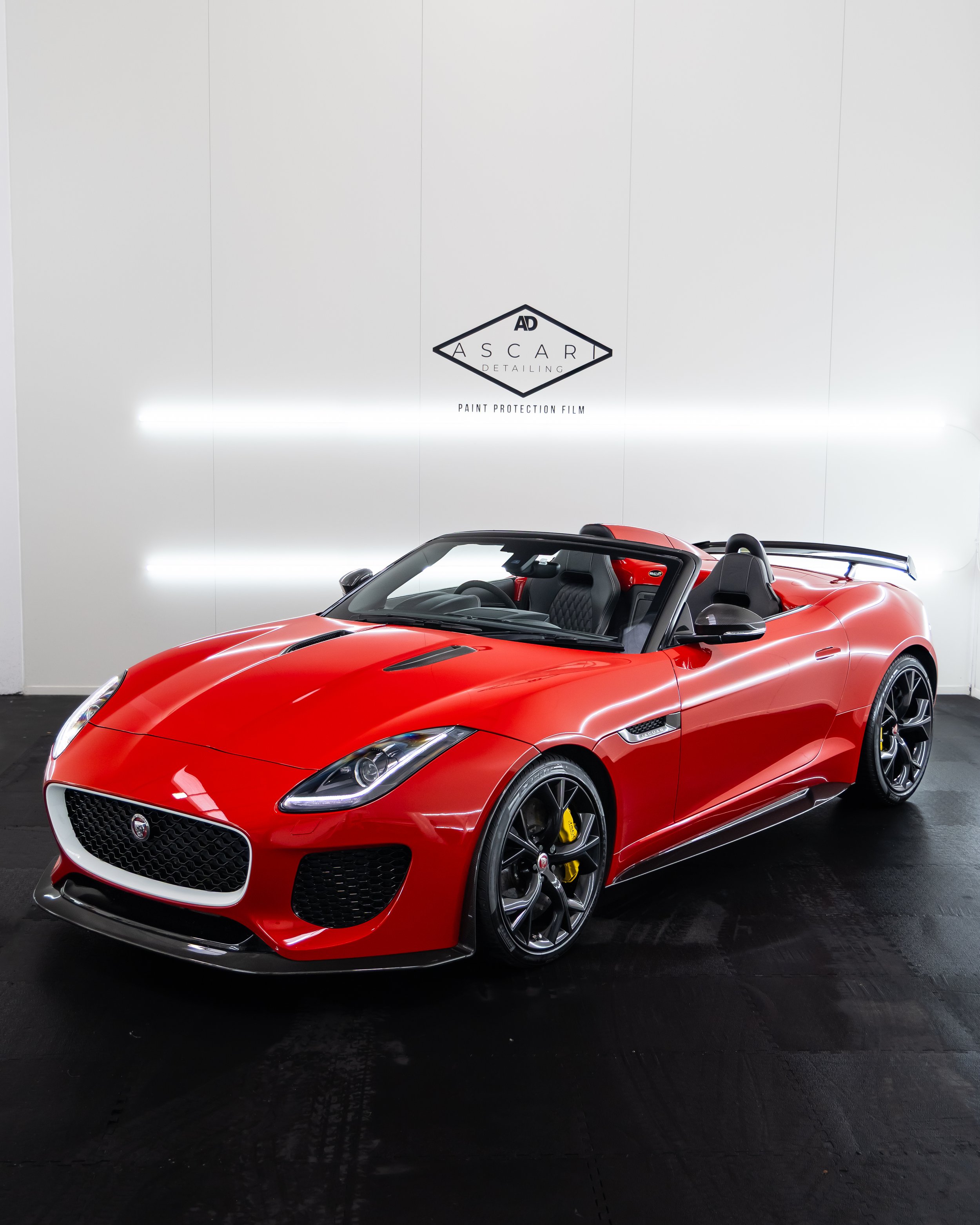 Red convertible sports car parked indoors with a white wall and black floor, featuring a sign that reads 'ASCARI Detailing' and 'Paint Protection Film'.
