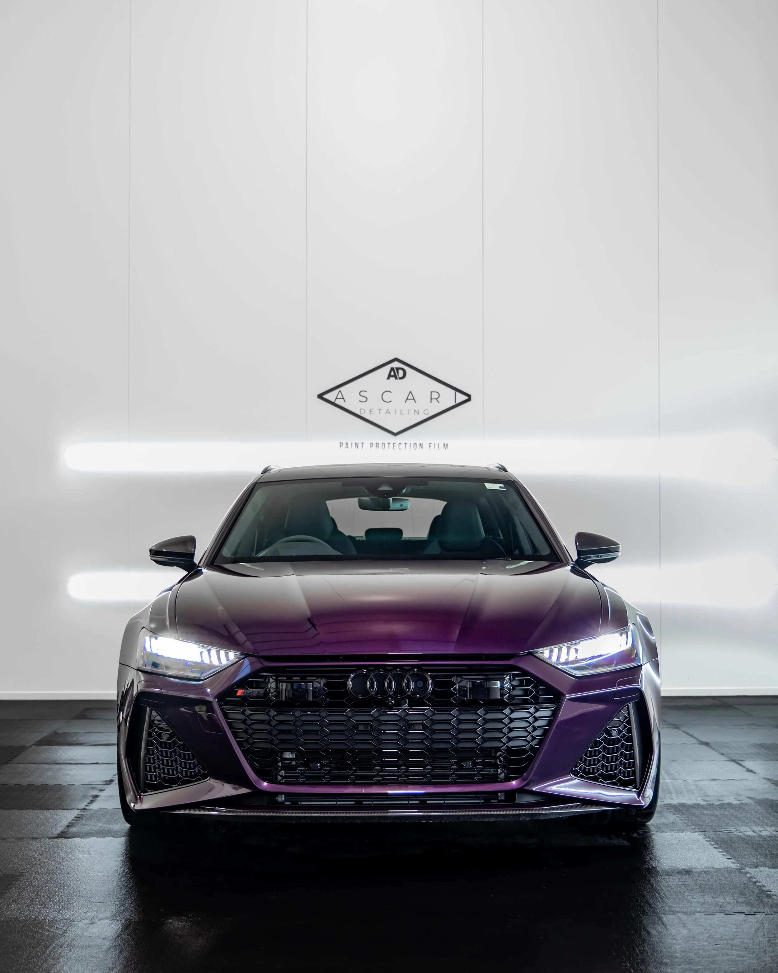 Front view of a dark purple Audi car with illuminated headlights, parked indoors against a white wall background with logo and text.