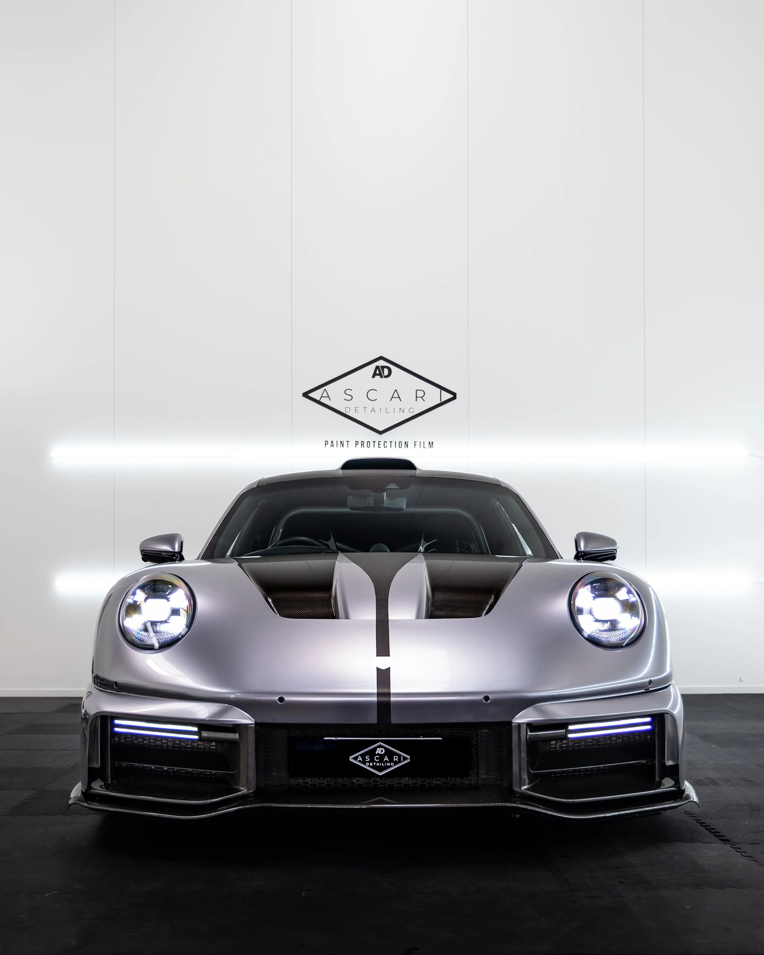 Modern silver sports car with black accents, front view in a detailing studio with white walls and LED lighting, branding for Ascari Detailing on the wall and license plate area.
