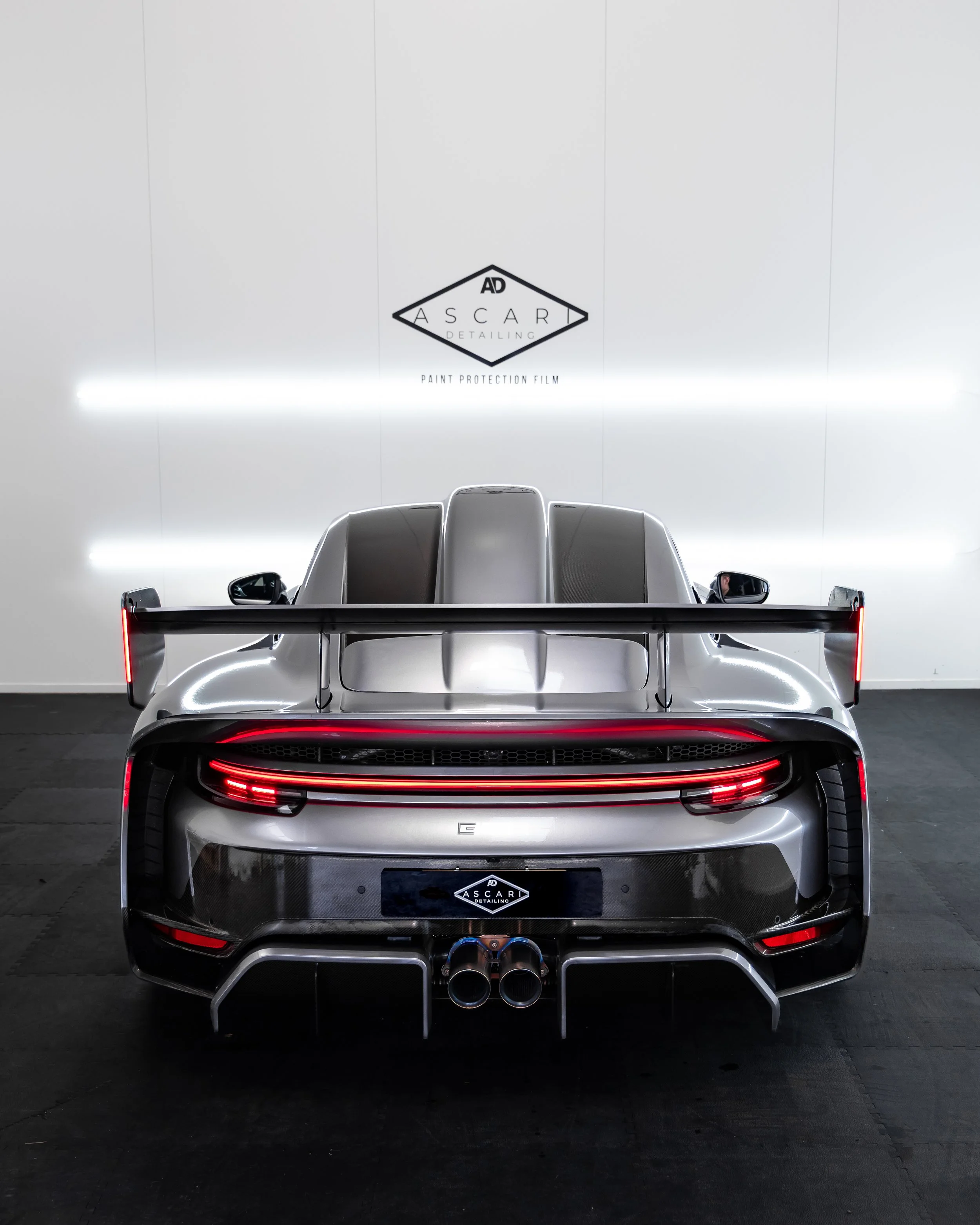 Rear view of a silver sports car with a large rear wing, featuring LED taillights, dual exhaust pipes, and a black diffuser, inside a showroom with a white wall and the logo 'Ascari Detailing'.