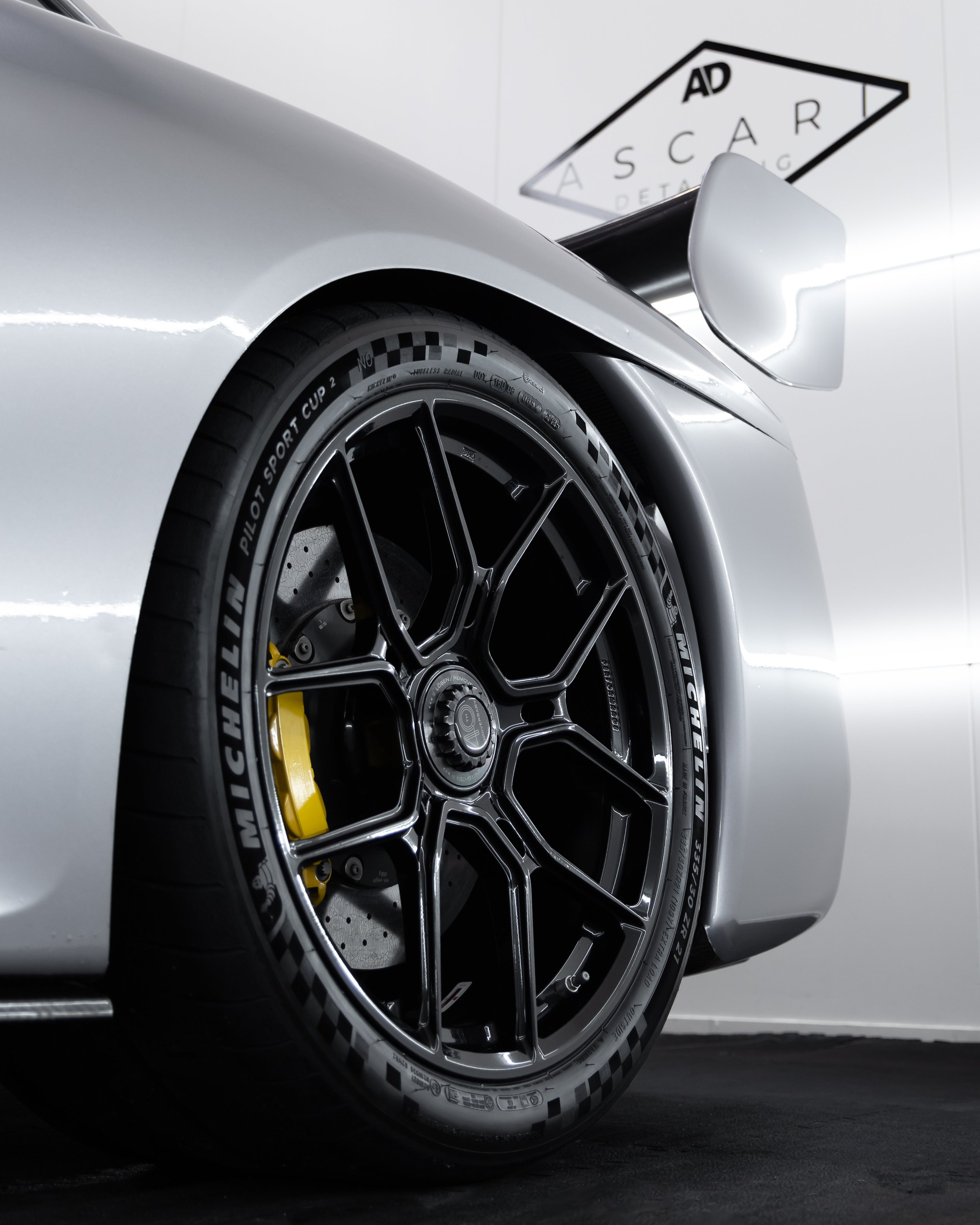 Close-up of a sleek silver race car with a black wheel featuring yellow brake calipers and Michelin tires, in a garage with a sign in the background.