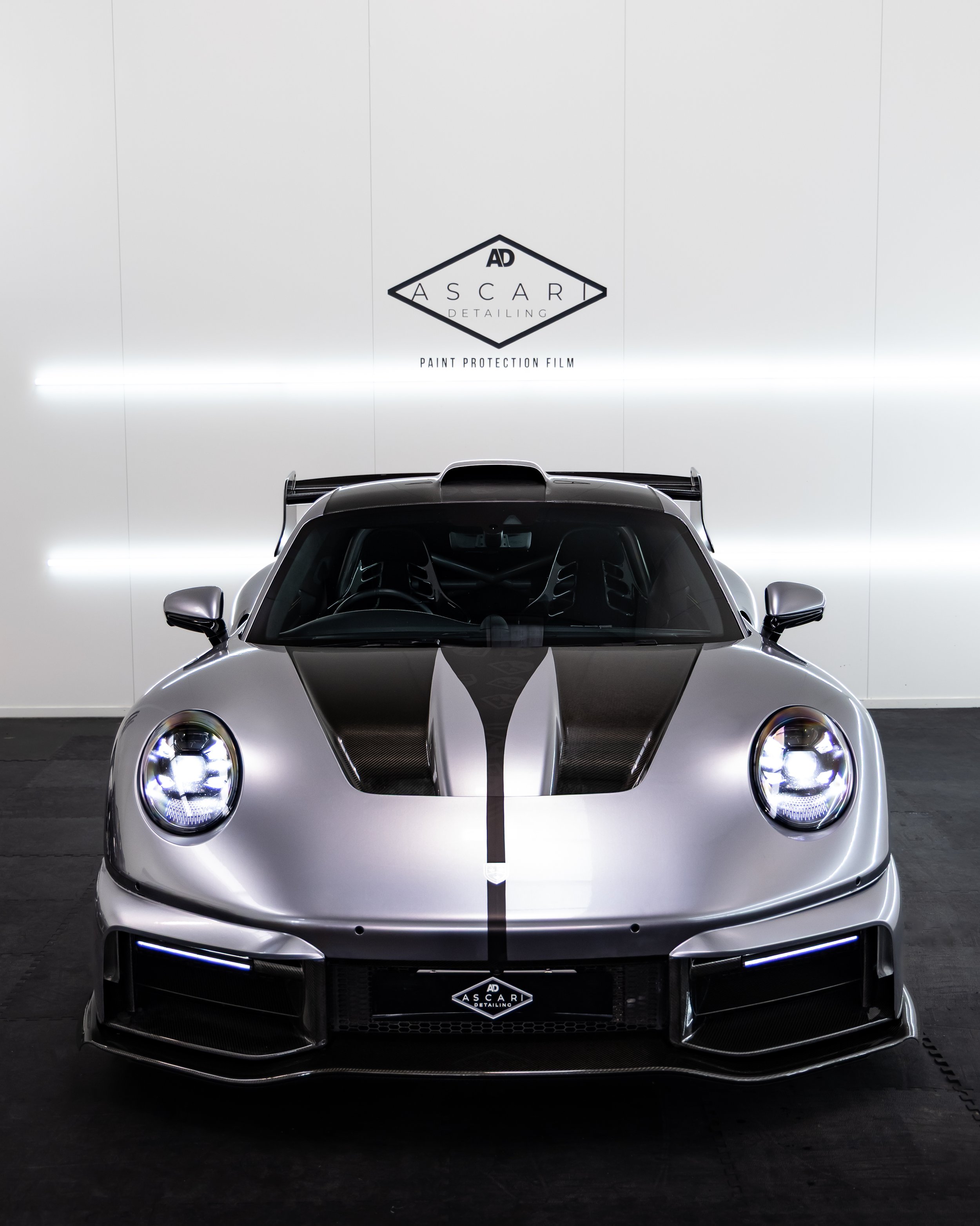 Silver sports car with black racing stripes, displayed indoors in front of a white wall with the Ascari Detailing logo and text.
