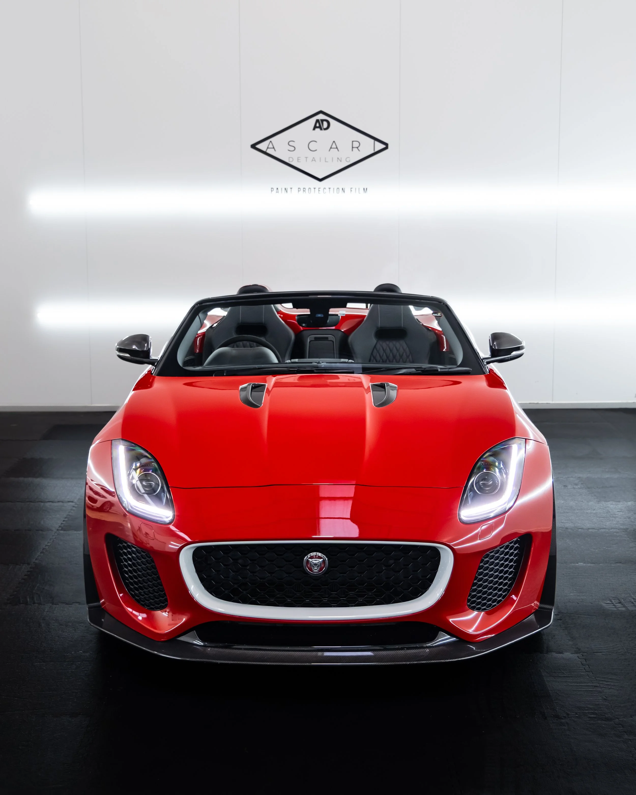 Red convertible sports car parked indoors against a white wall with a sign for 'ASCARI Detailing' and paint protection film.