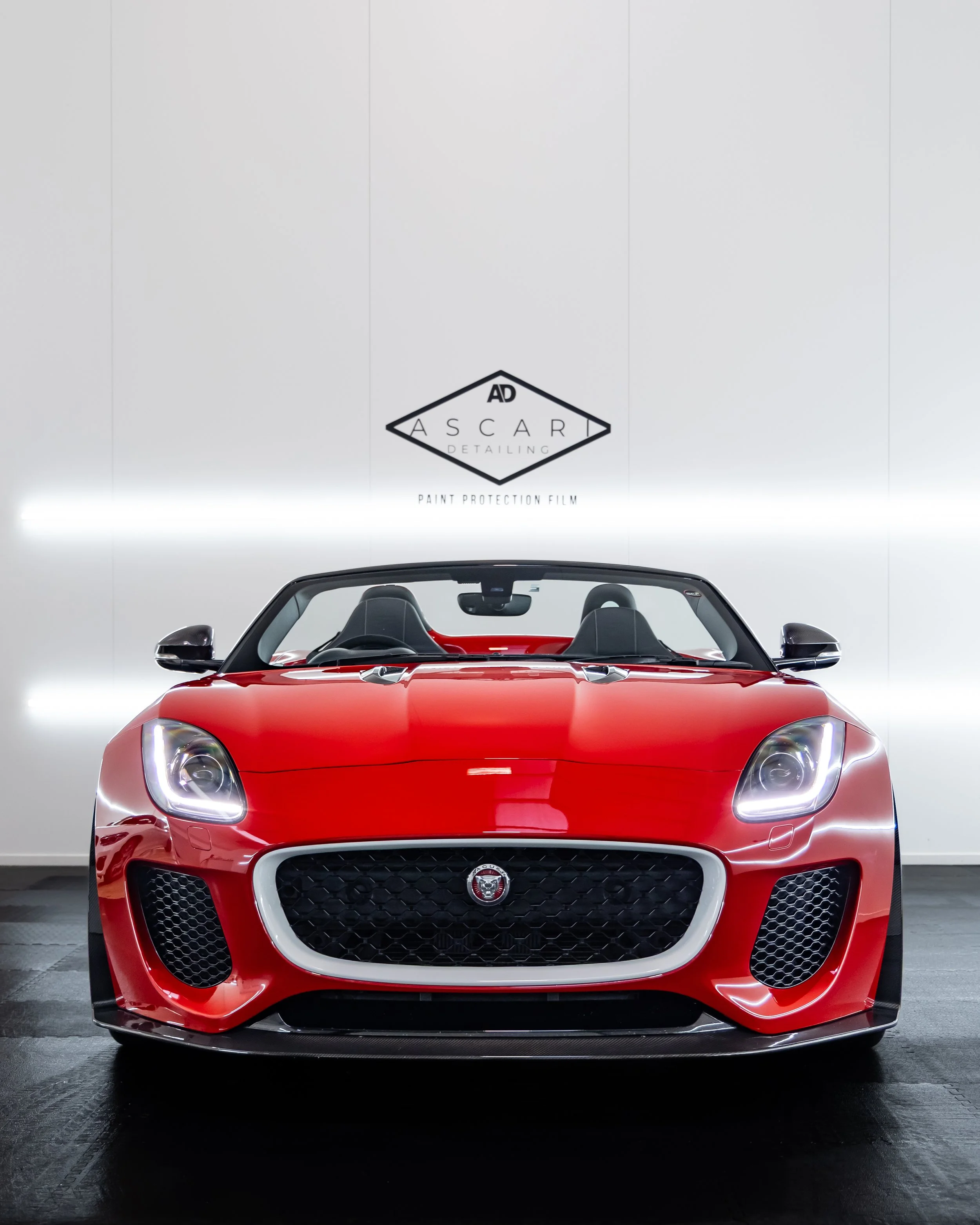Red sports car with open top inside a showroom, with a white wall and signage in the background that reads 'ASCARI Detailing' and 'Paint Protection Film'.