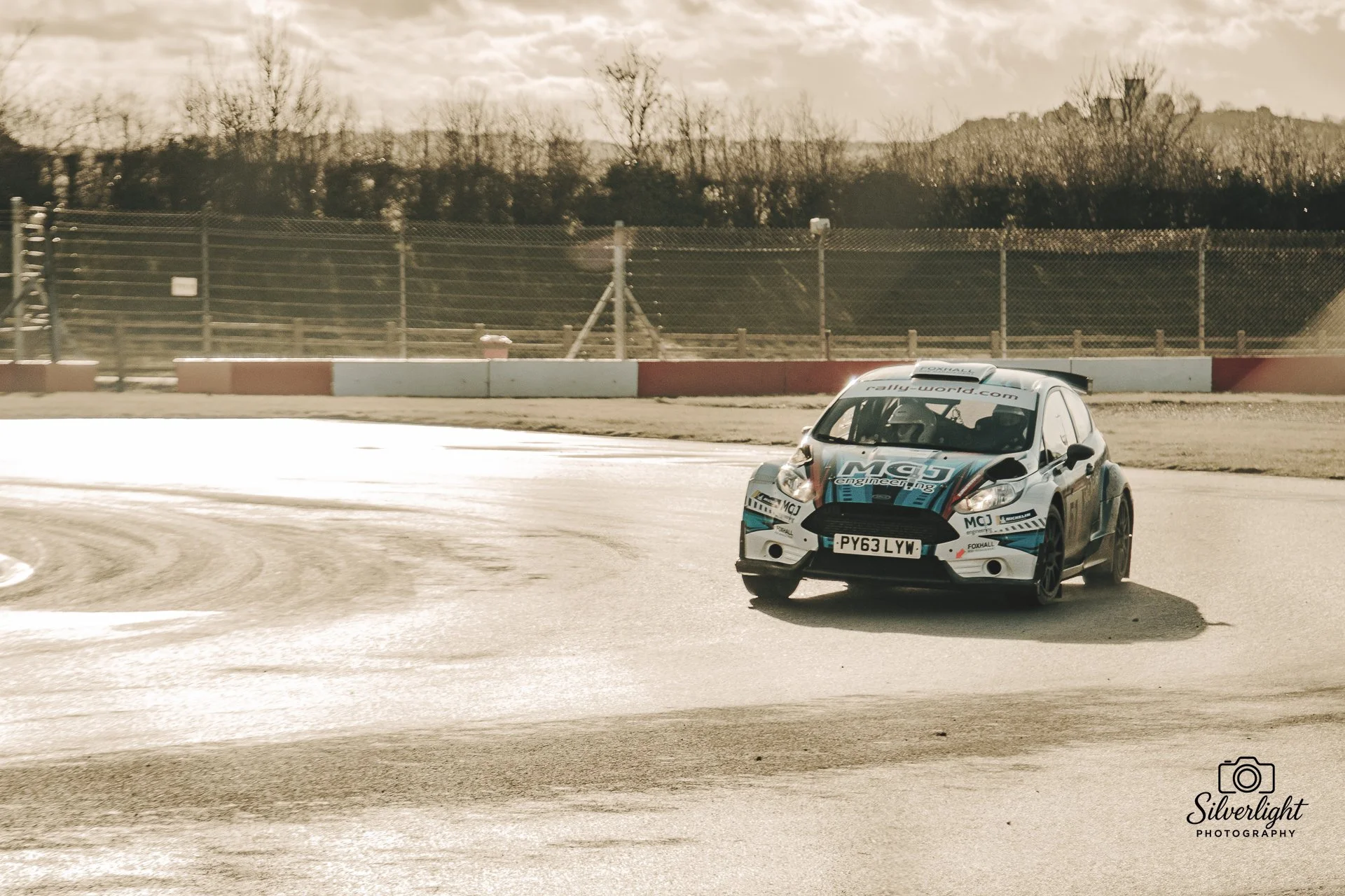 A rally car on at donington race track during daylight, with a fence and trees in the background.