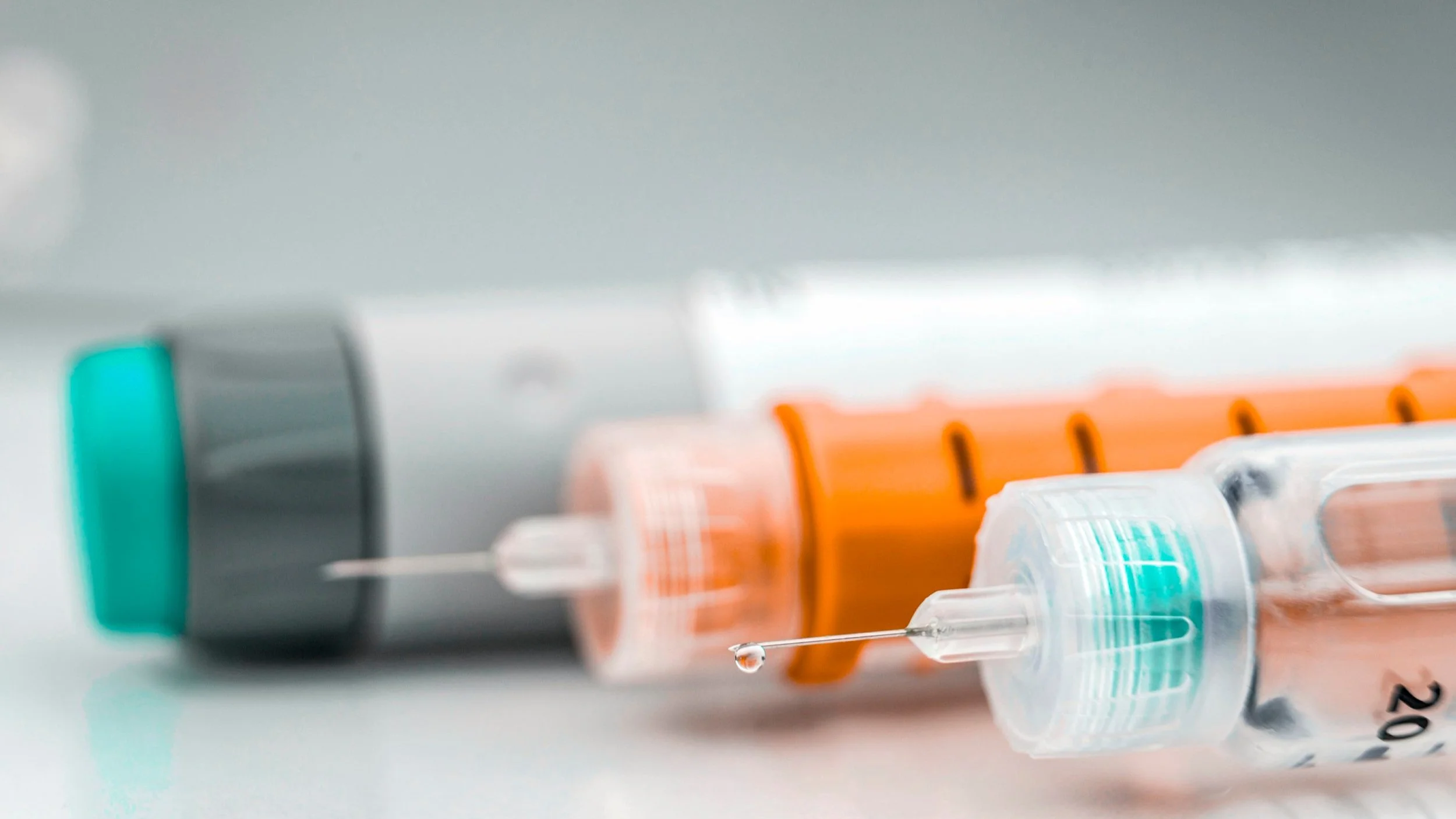 Close-up of two syringes with orange and gray caps, one with a drop of liquid on the needle, lying on a white surface.