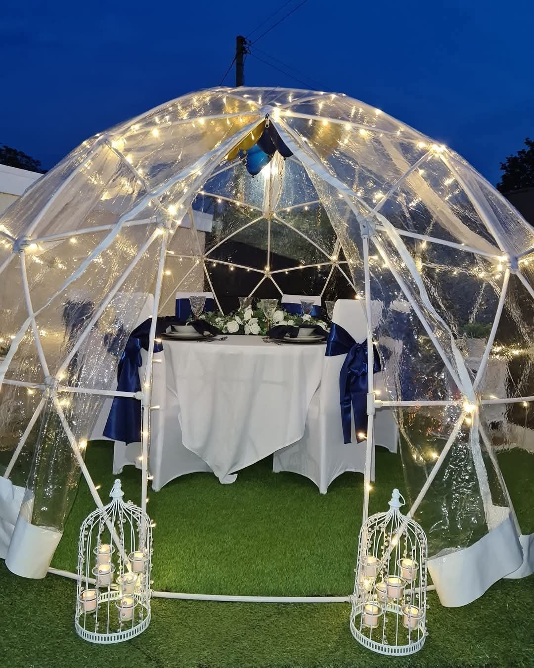 A nighttime outdoor dining setup inside a transparent geodesic dome decorated with string lights, with a table set with black and white tableware, and surrounded by white birdcage-shaped candle holders with candles.
