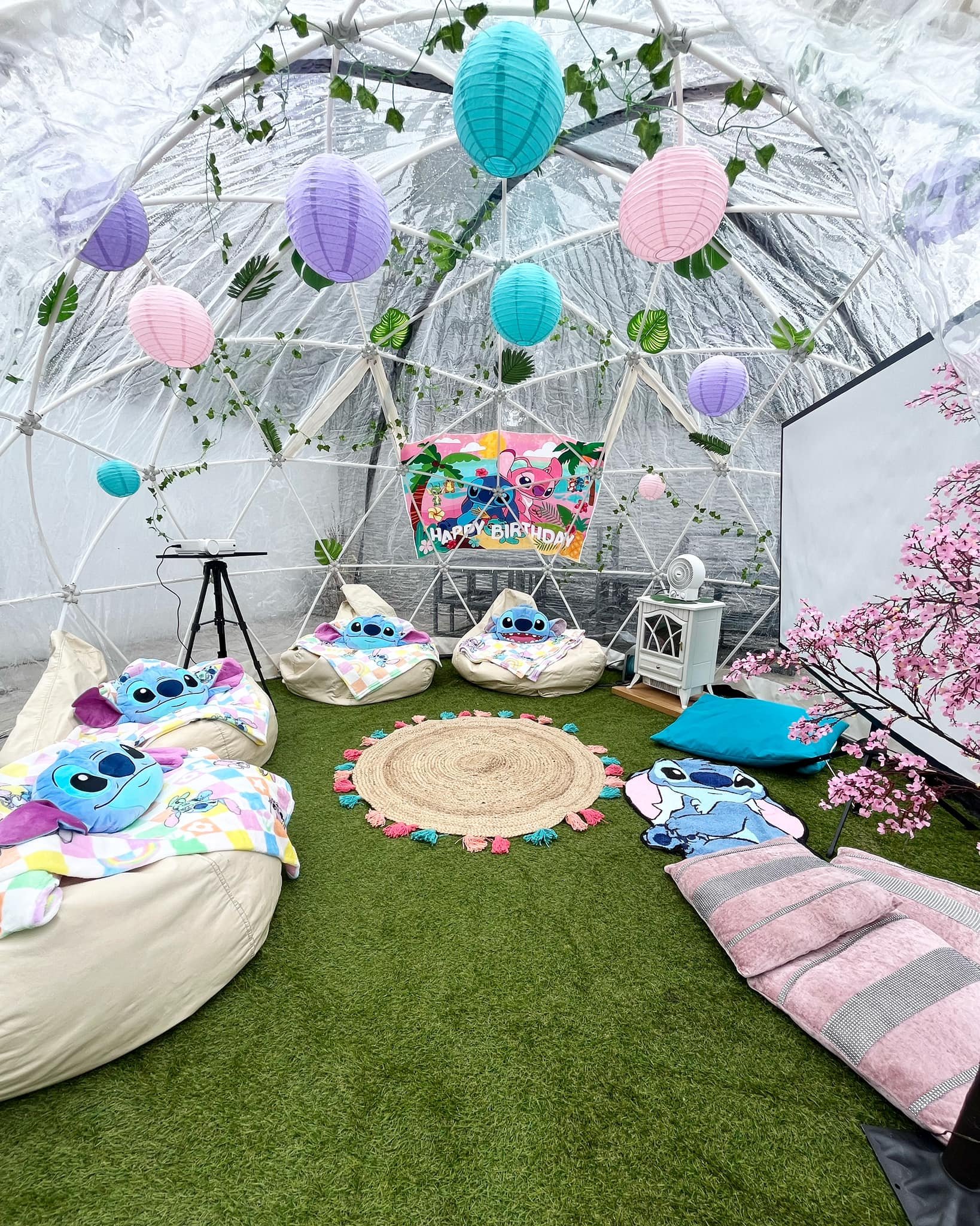 Indoor space decorated for a birthday party with Stitch from Lilo & Stitch theme, featuring hanging paper lanterns in pink, purple, and blue, green vines, a 'Happy Birthday' banner with Stitch and tropical scenery, plush Stitch pillows on bean bag ch