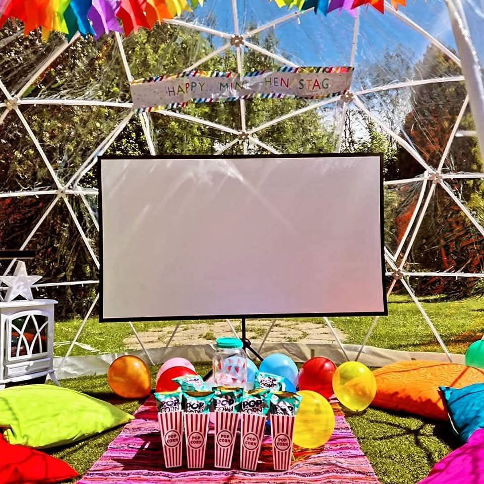 Outdoor party setup with a geodesic dome, colorful balloons, popcorn boxes, pillows, and a large white screen for a movie or presentation, decorated with a banner reading 'Happy Minkenstag.'
