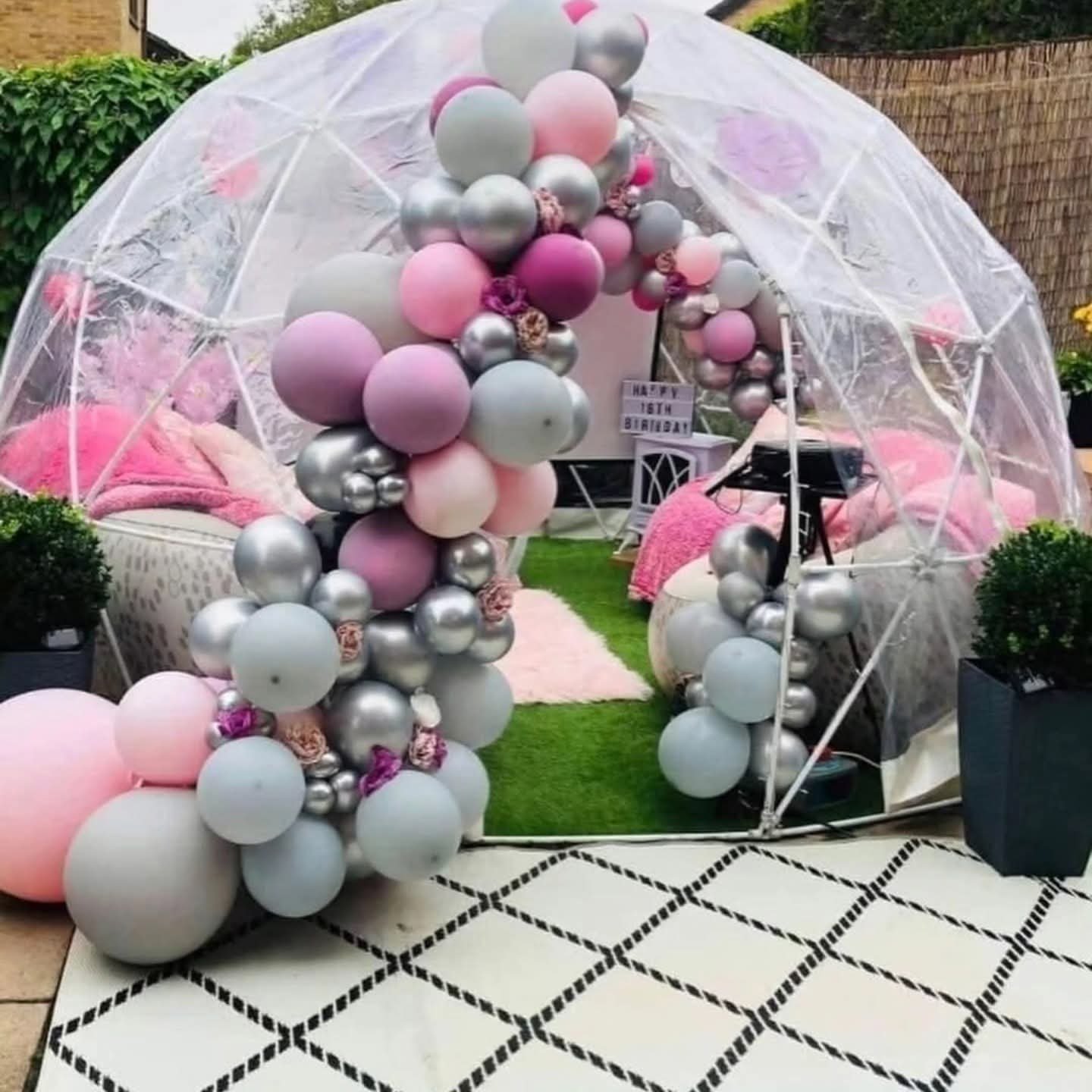 Decorated outdoor space with a transparent geodesic dome, pink and gray balloons, plush cushions, and a sign that reads "Happy 18th Birthday."