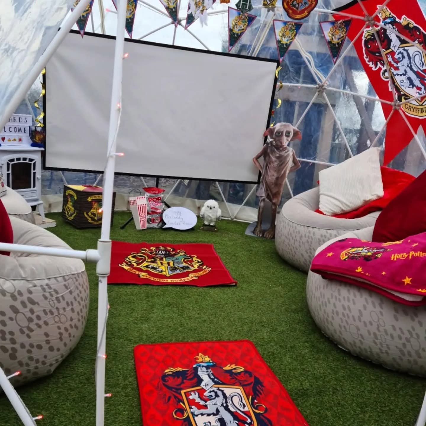 A Harry Potter themed indoor space with Harry Potter blankets, banners, and furniture, featuring a life-sized dog mascot, a small plush owl, a popcorn container, and a sign that says '#1 Bulldog Fan Place,' inside a tent with transparent sides.