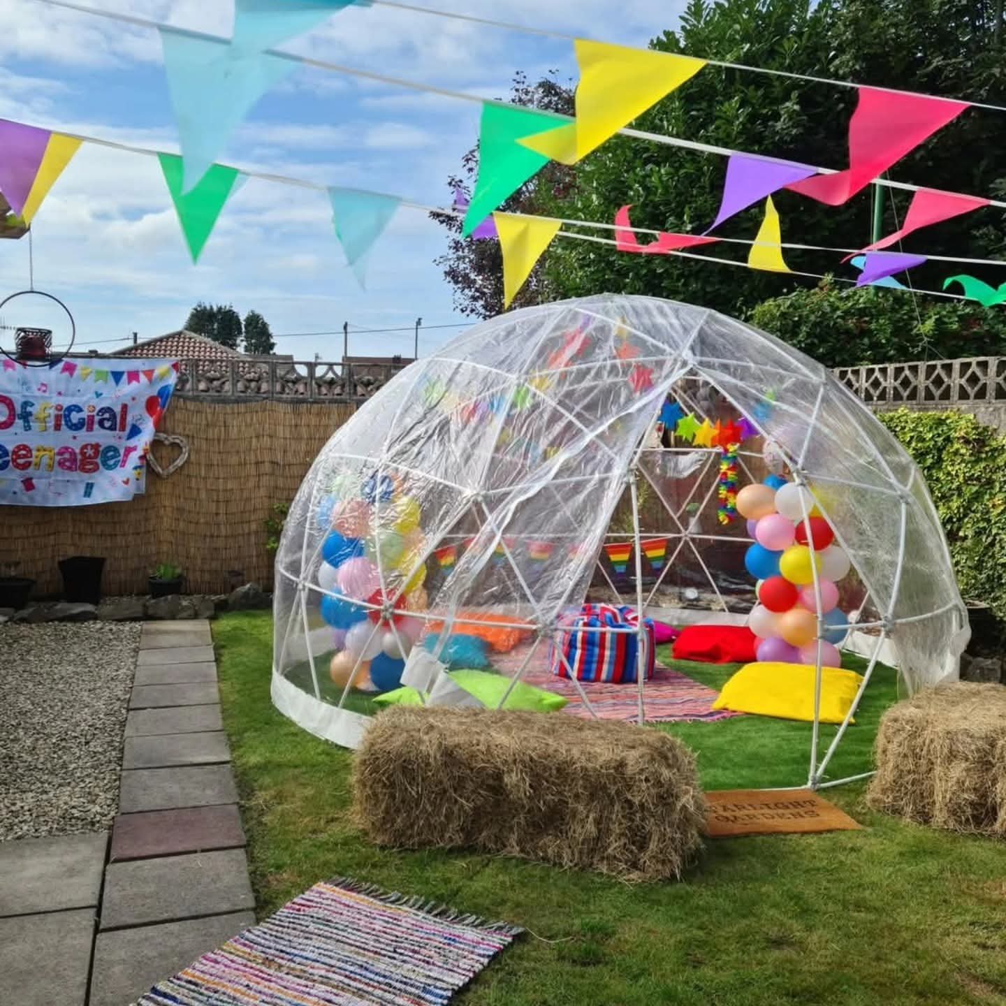 Outdoor birthday party setup with a clear geodesic dome filled with balloons and colorful cushions, adorned with rainbow banners and buntings, on a grassy backyard.