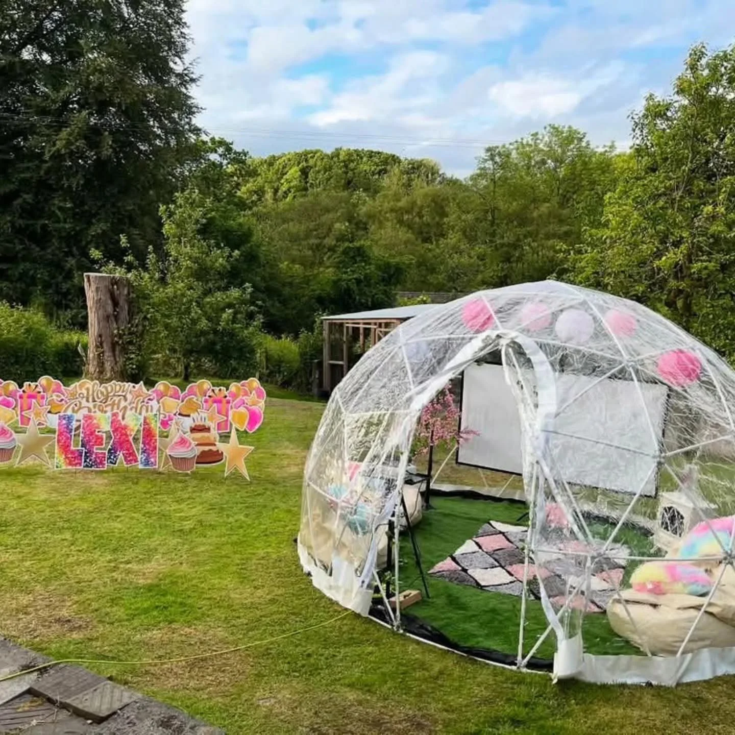 Outdoor birthday celebration setup with a clear tent decorated with pink and white balloons, a sign that reads 'Happy Birthday LEXI', and a grassy area with trees and a cloudy sky.