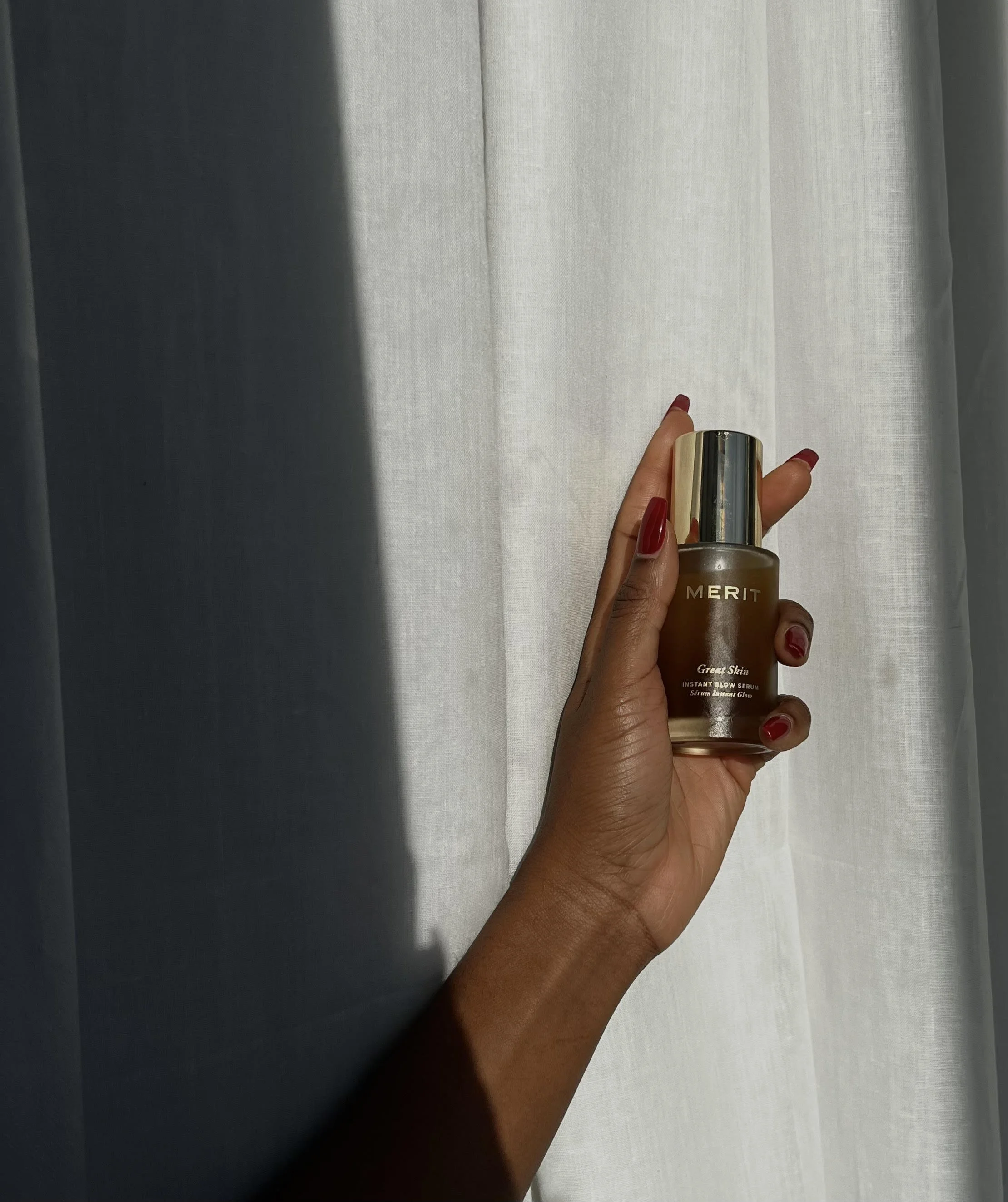 A hand holding a bottle of Merit Great Skin Instant Glow Serum in front of light-colored curtains.