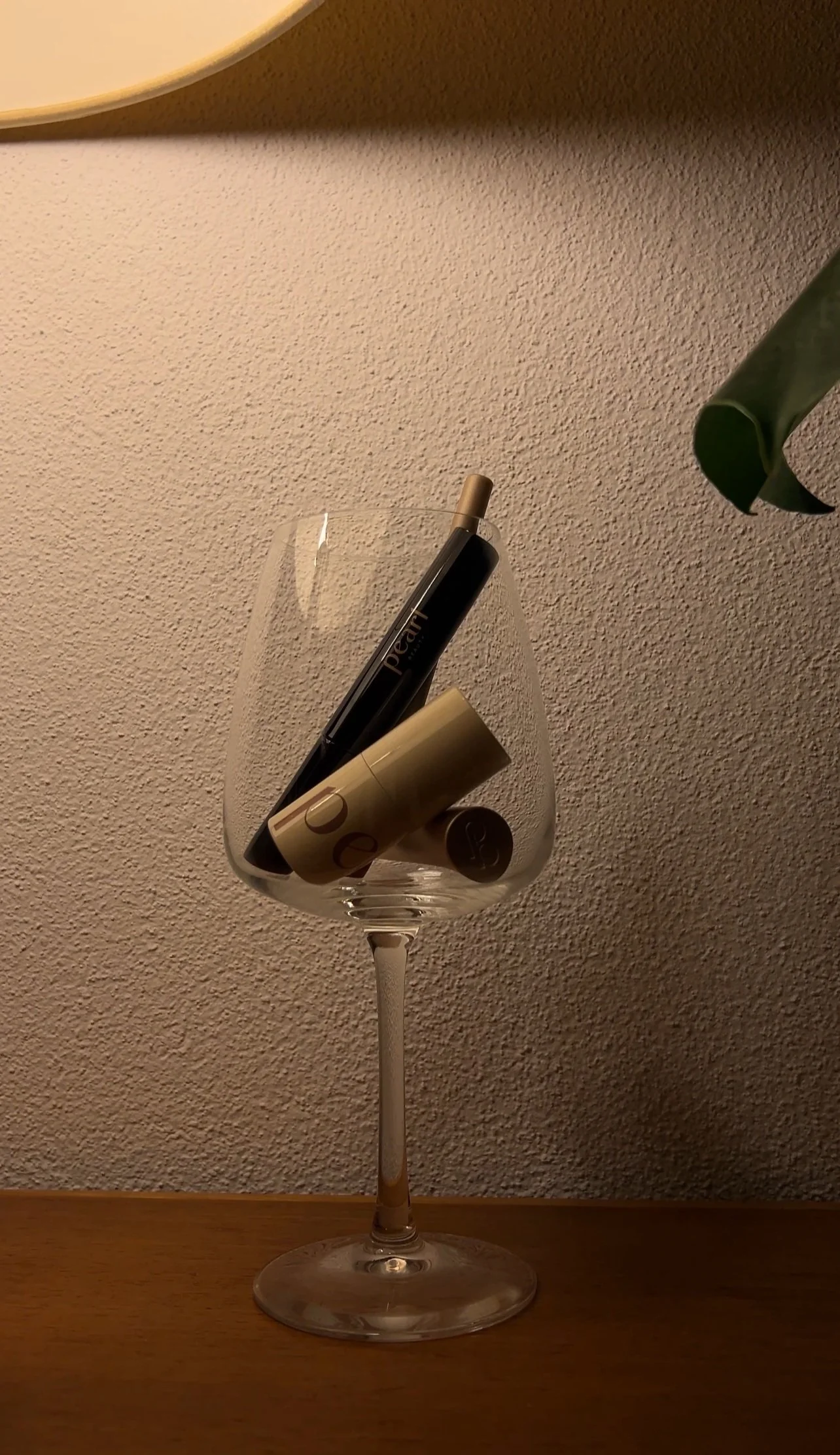 A clear wine glass holding two lip balm sticks, one black and one gold, on a wooden surface against a textured beige wall.