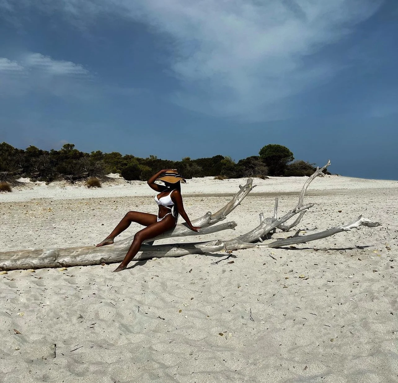 A woman in a white bikini sitting on a large piece of driftwood on a sandy beach under a partly cloudy sky.