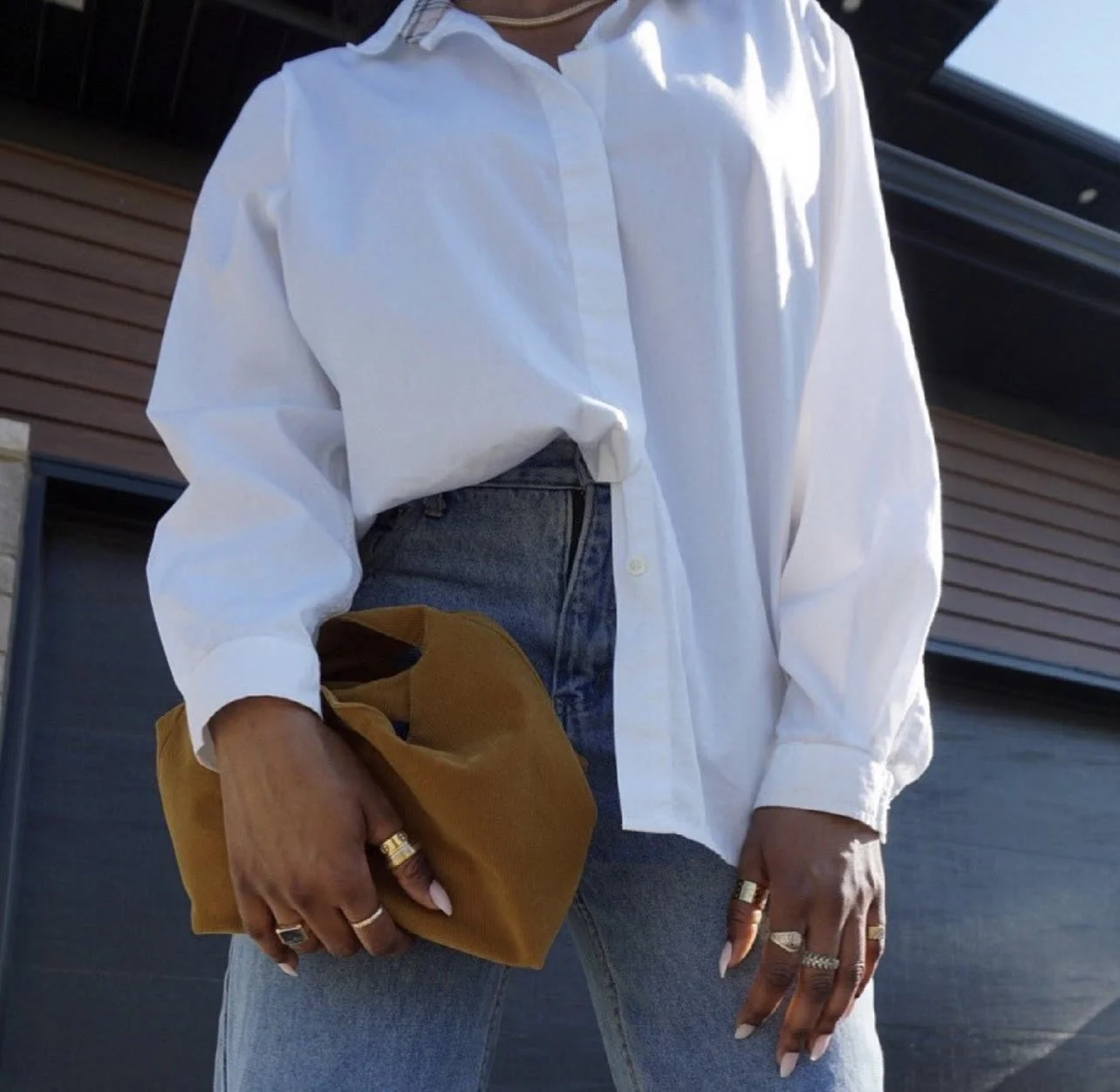 Person wearing a white button-up shirt tucked into high-waisted blue jeans, holding a brown handbag, with multiple rings on their fingers.