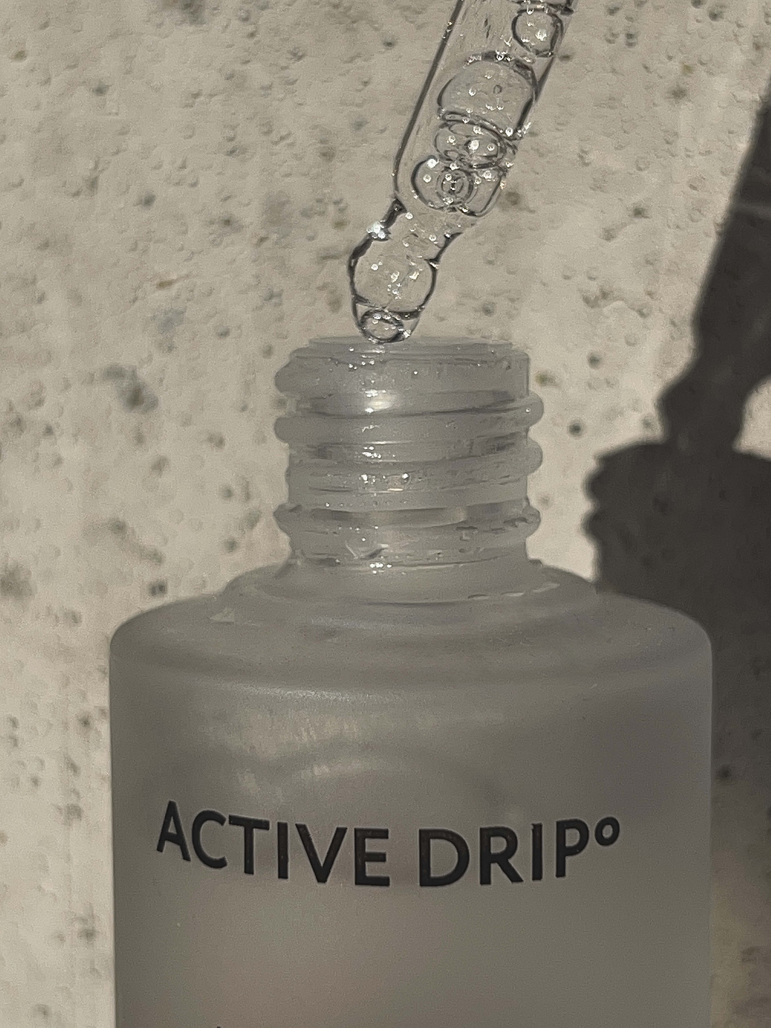 Close-up of a clear dropper releasing liquid into a frosted glass bottle labeled "ACTIVE DRIP" with a textured light-colored surface in the background.