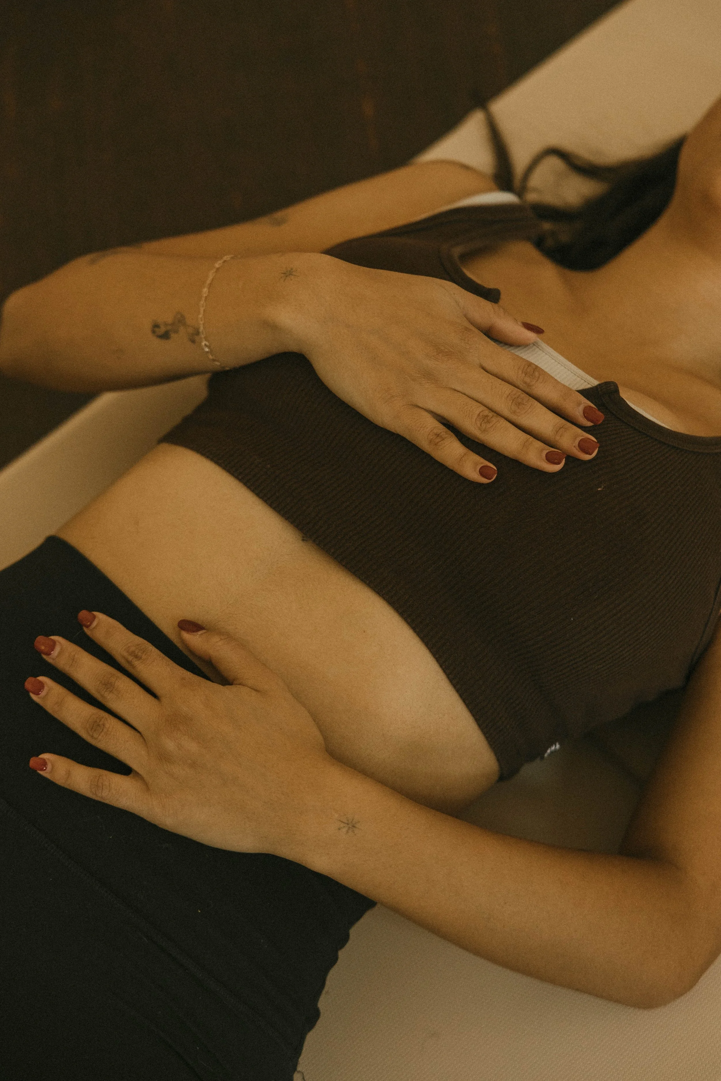 A woman lying down, wearing a black crop top and black pants, with hands resting on her chest and stomach. She has dark hair, painted nails, jewelry, and tattoos on her arms.