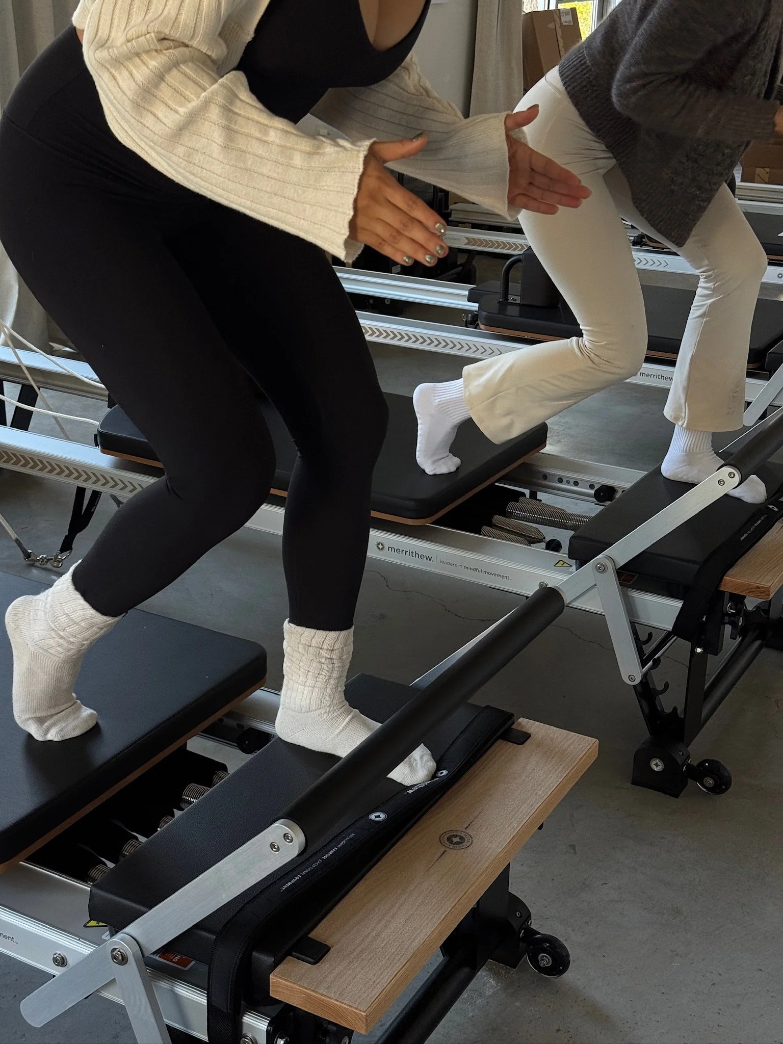 Two women exercising on Pilates reformers, demonstrating squats, in a fitness studio.
