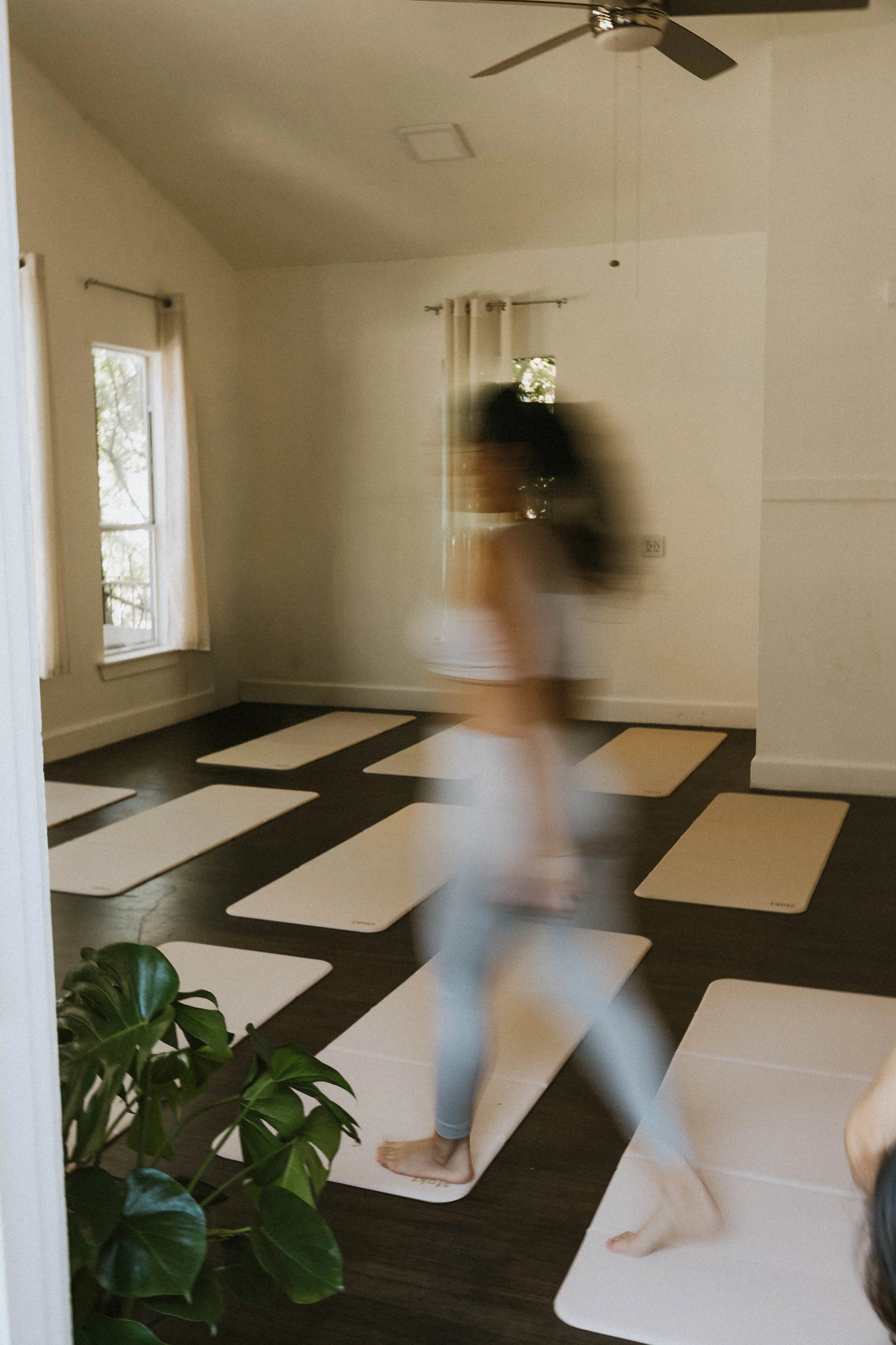 A person practicing yoga on a white mat in a bright, minimalistic room with dark wooden floors, windows, and a ceiling fan.