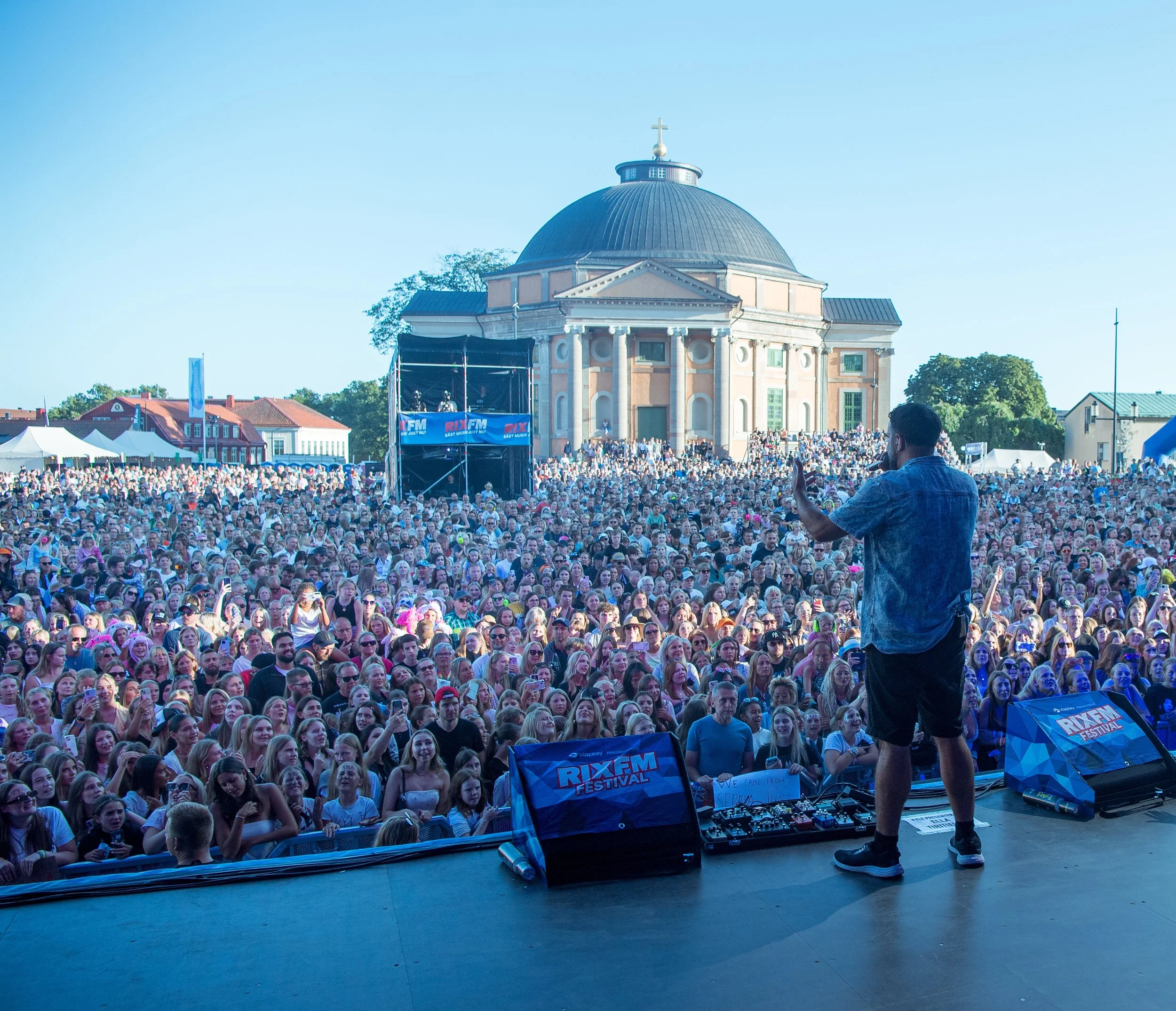 A large outdoor concert with Simon Lingmerth performing on stage and a crowd of people enjoying the music. In the background, there is a historic building with a dome and cross, and blue skies.