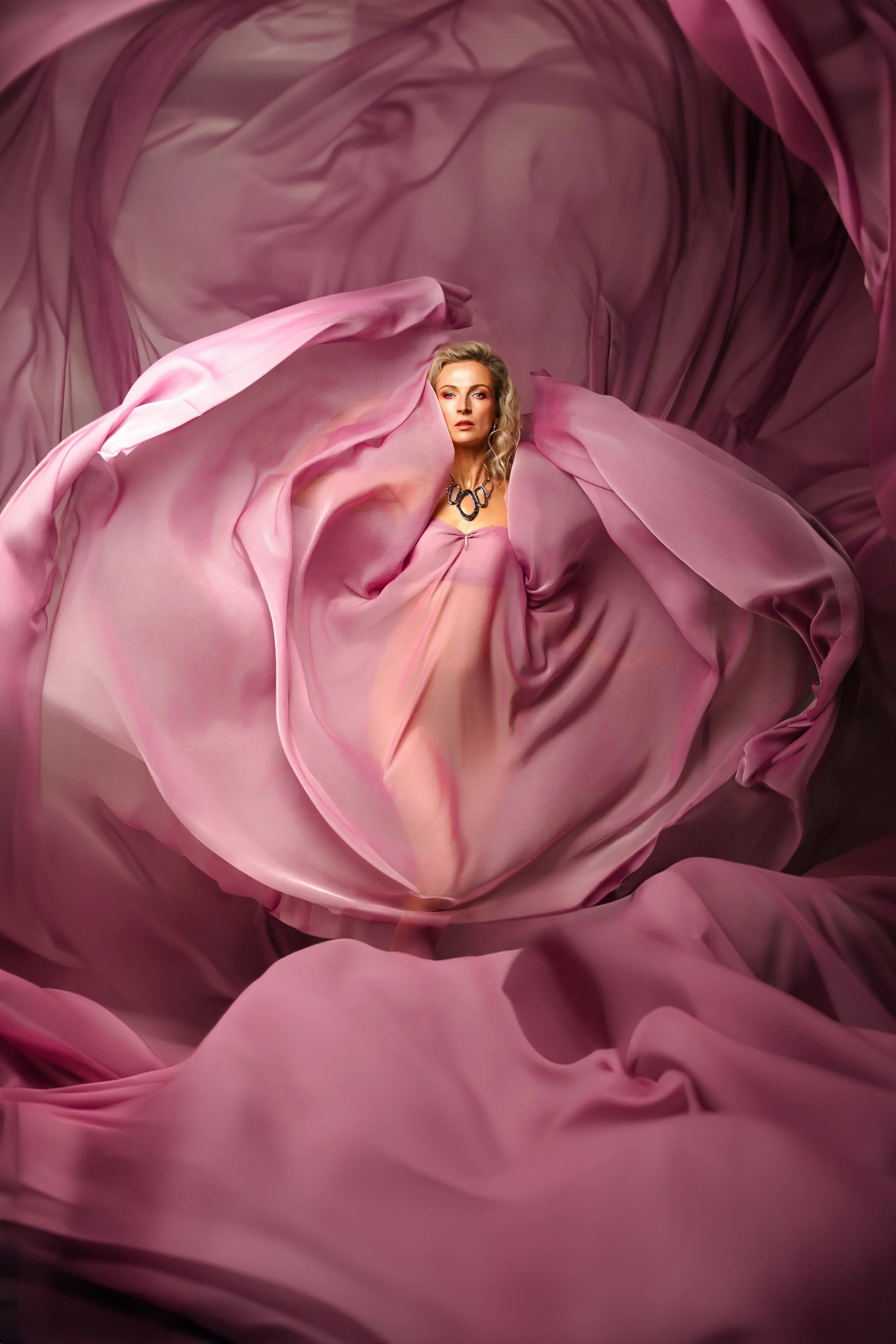 A woman with blonde hair wearing a pink dress and a black necklace, surrounded by flowing pink and purple fabric that creates a swirling, abstract background.