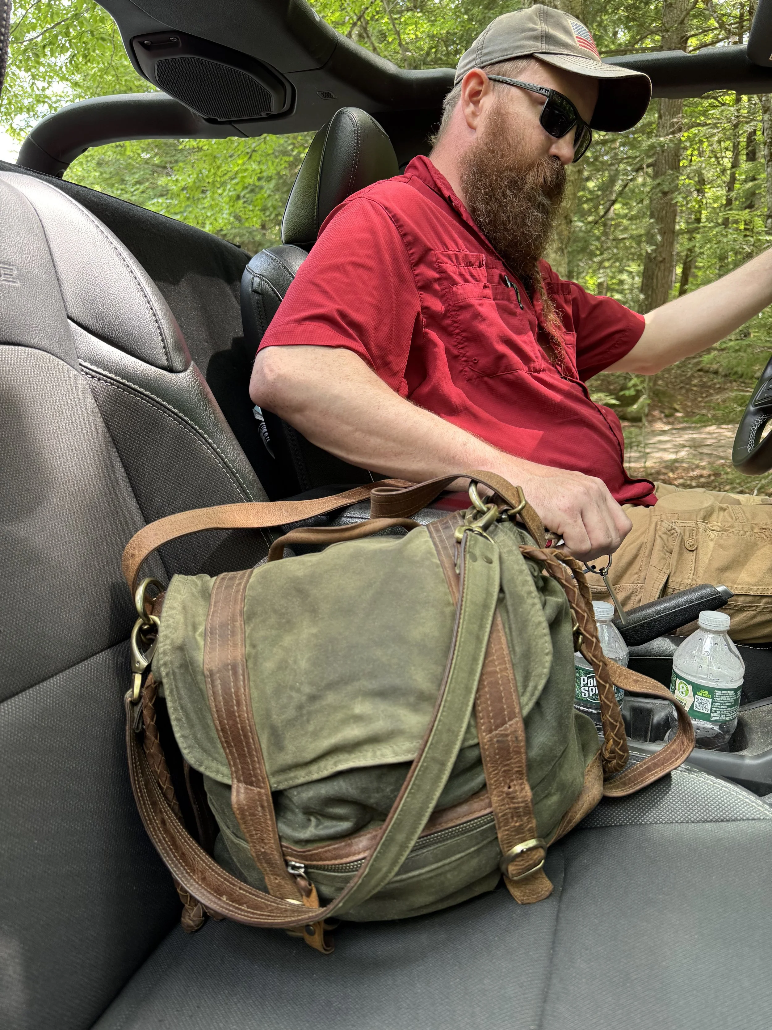 Joshua, wearing sunglasses, a cap with an American flag, and a red shirt, is sitting in a vehicle in a forested area. The Stonington pack sits on the seat in front of him.
