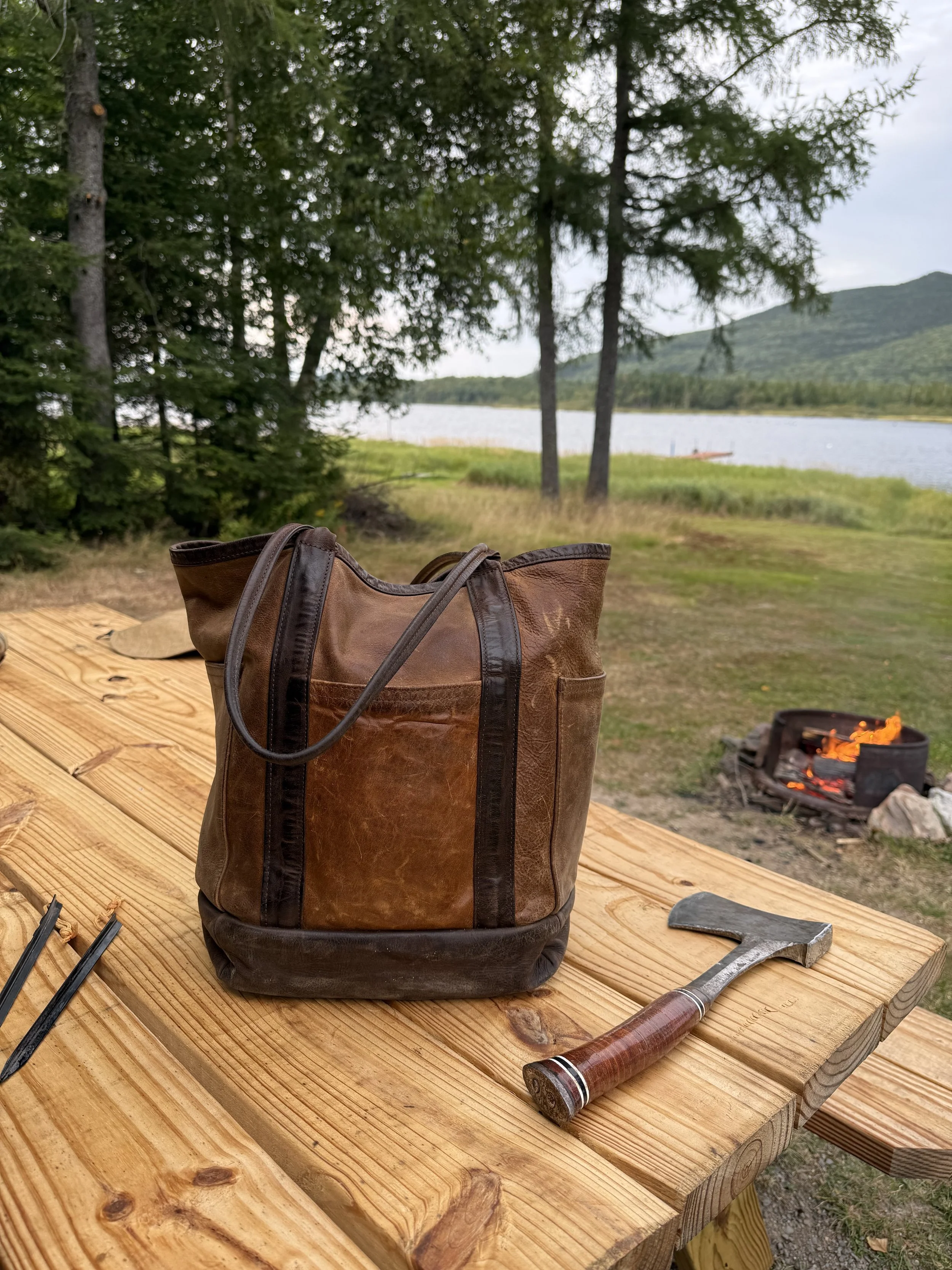 Kingfield on a wooden table with a hammer, tongs, and a fire pit nearby, set outdoors near a lake with trees and hills in the background.