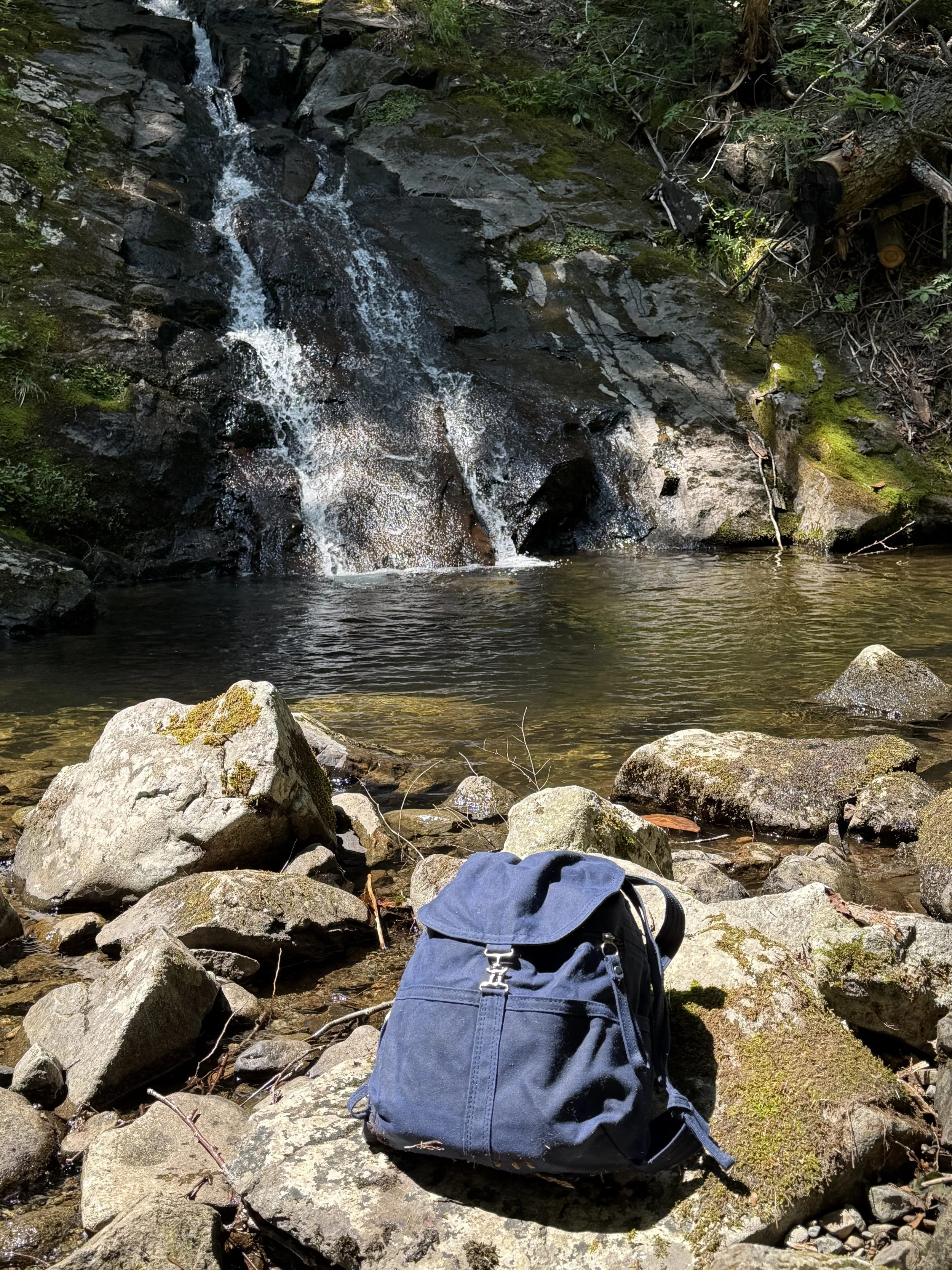 A small waterfall flowing over rocks into a calm pond surrounded by rocks and green foliage, with a blue canvas Rangeley backpack at the bottom of the image.