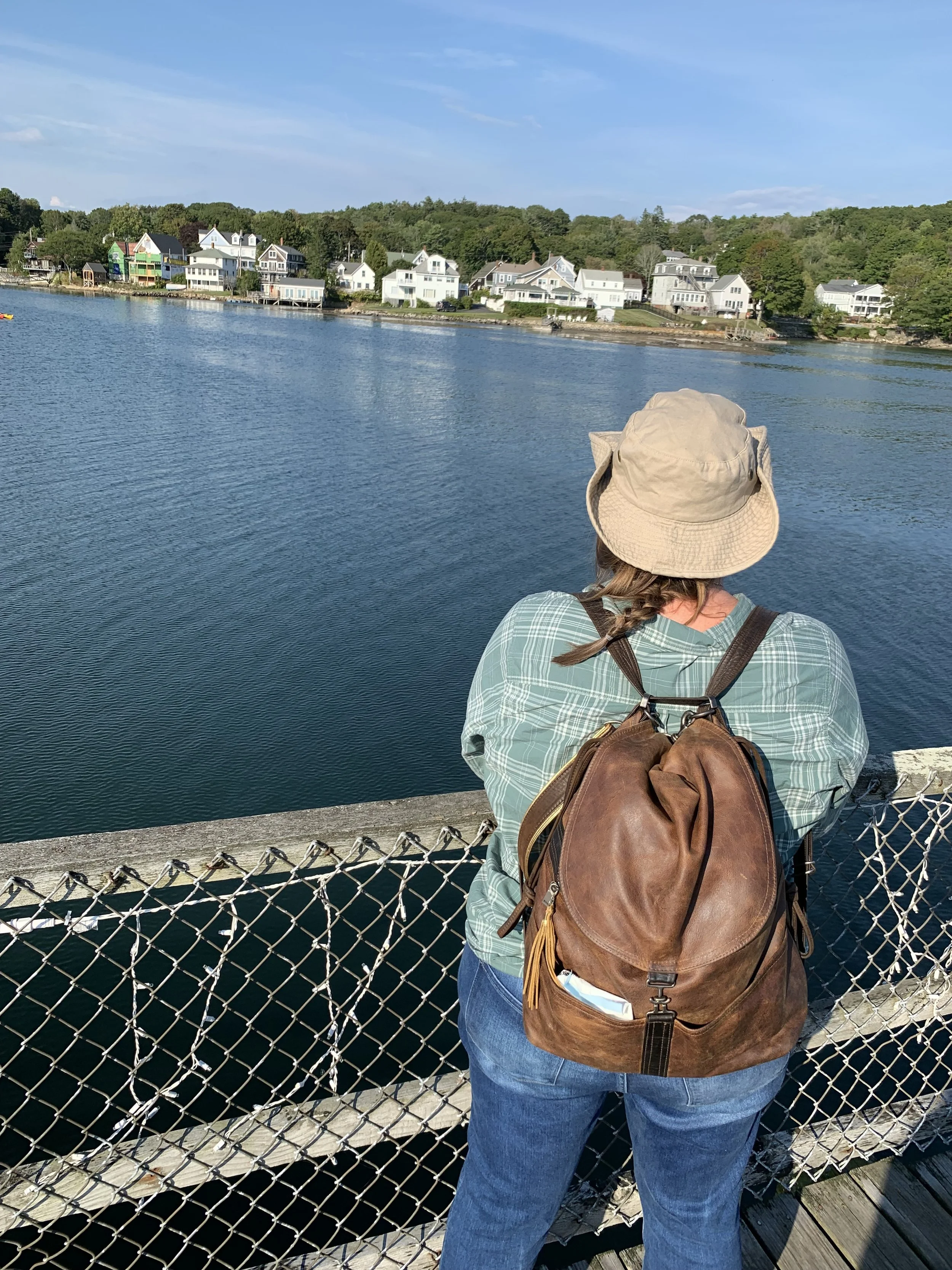 Jennifer with her Rangeley and wide-brimmed tan hat in Boothbay Harbor leaning on a waterfront fence, overlooking calm blue water and houses on a hill in the distance.